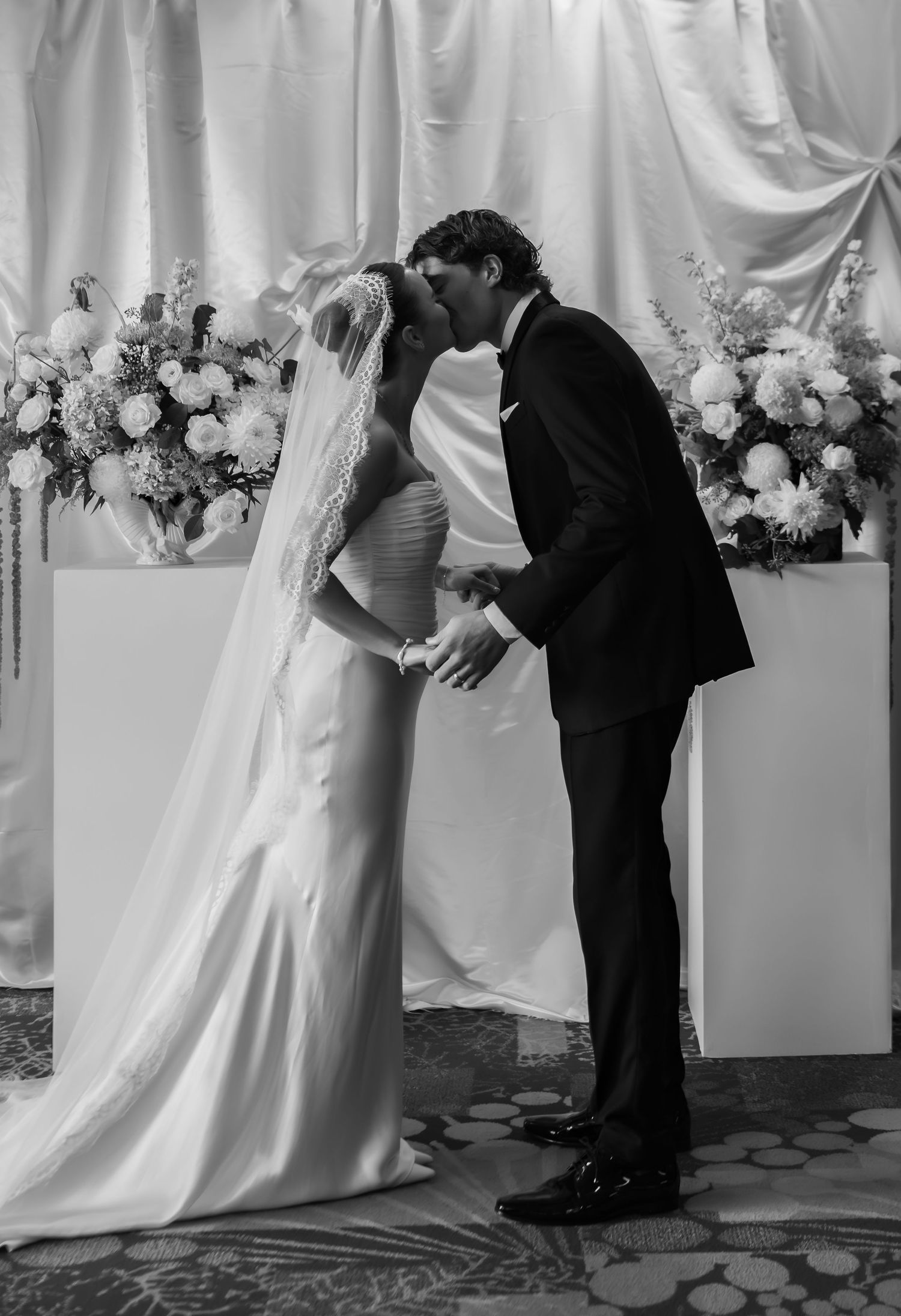 Black and white photo of a bride and groom sharing their first kiss at the wedding altar with floral arrangements.