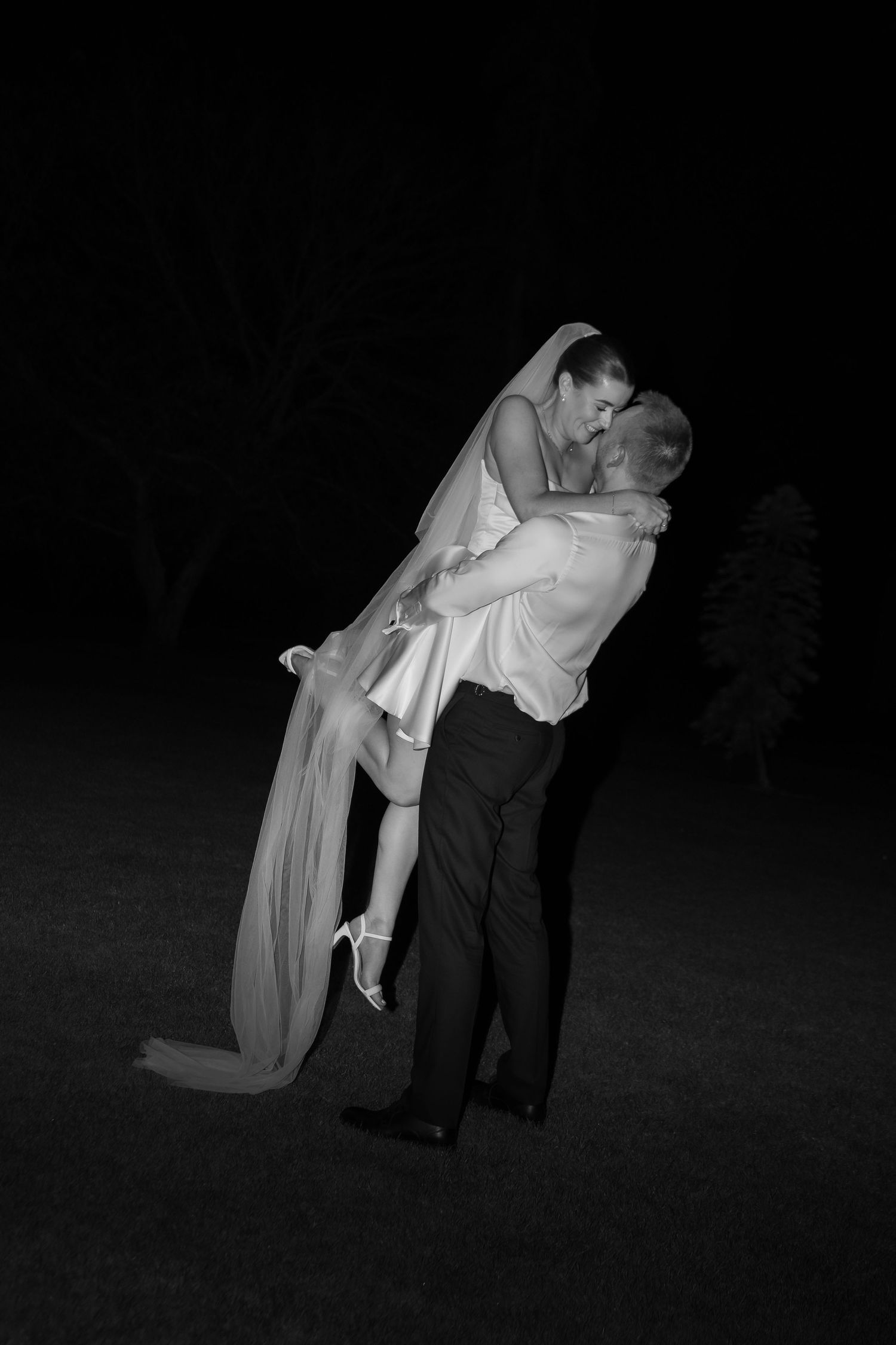 Groom lifting bride in wedding dress and veil, sharing romantic kiss outdoors at night in black and white photo.