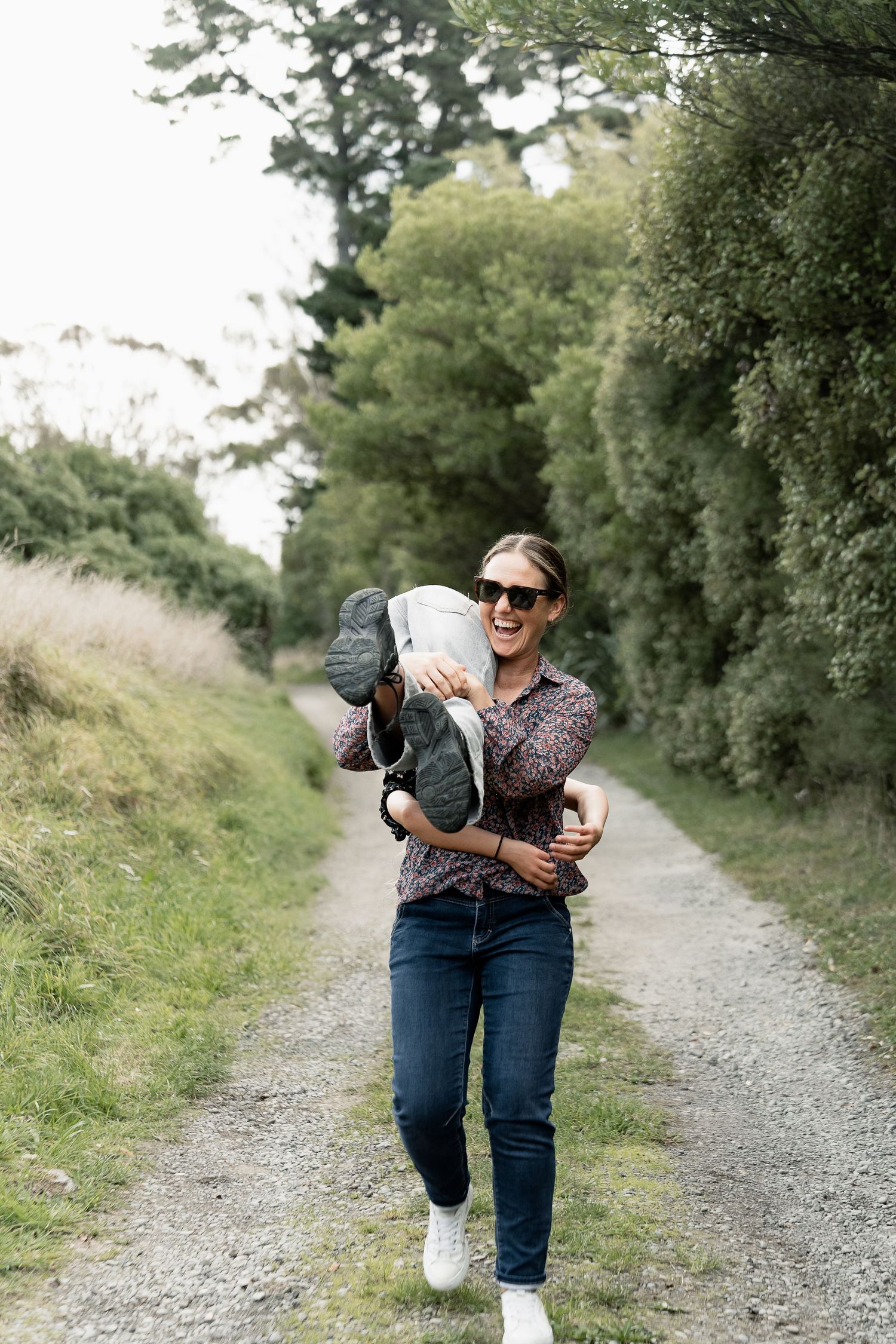 A smiling woman in sunglasses carries a child on a scenic gravel path surrounded by lush green trees.