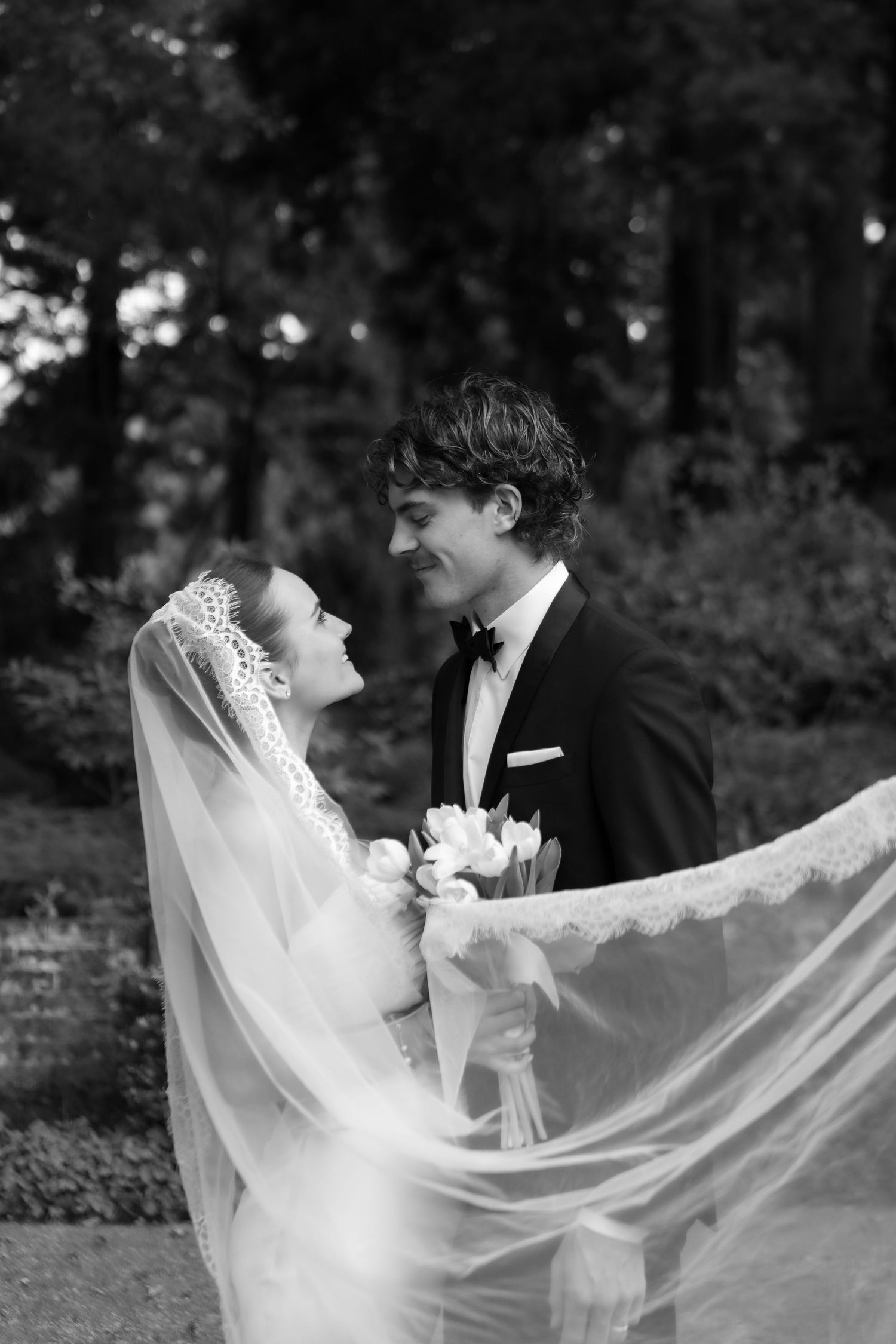 Black and white wedding photo of new zealand bride in veil and groom in tuxedo gazing at each other in a garden.