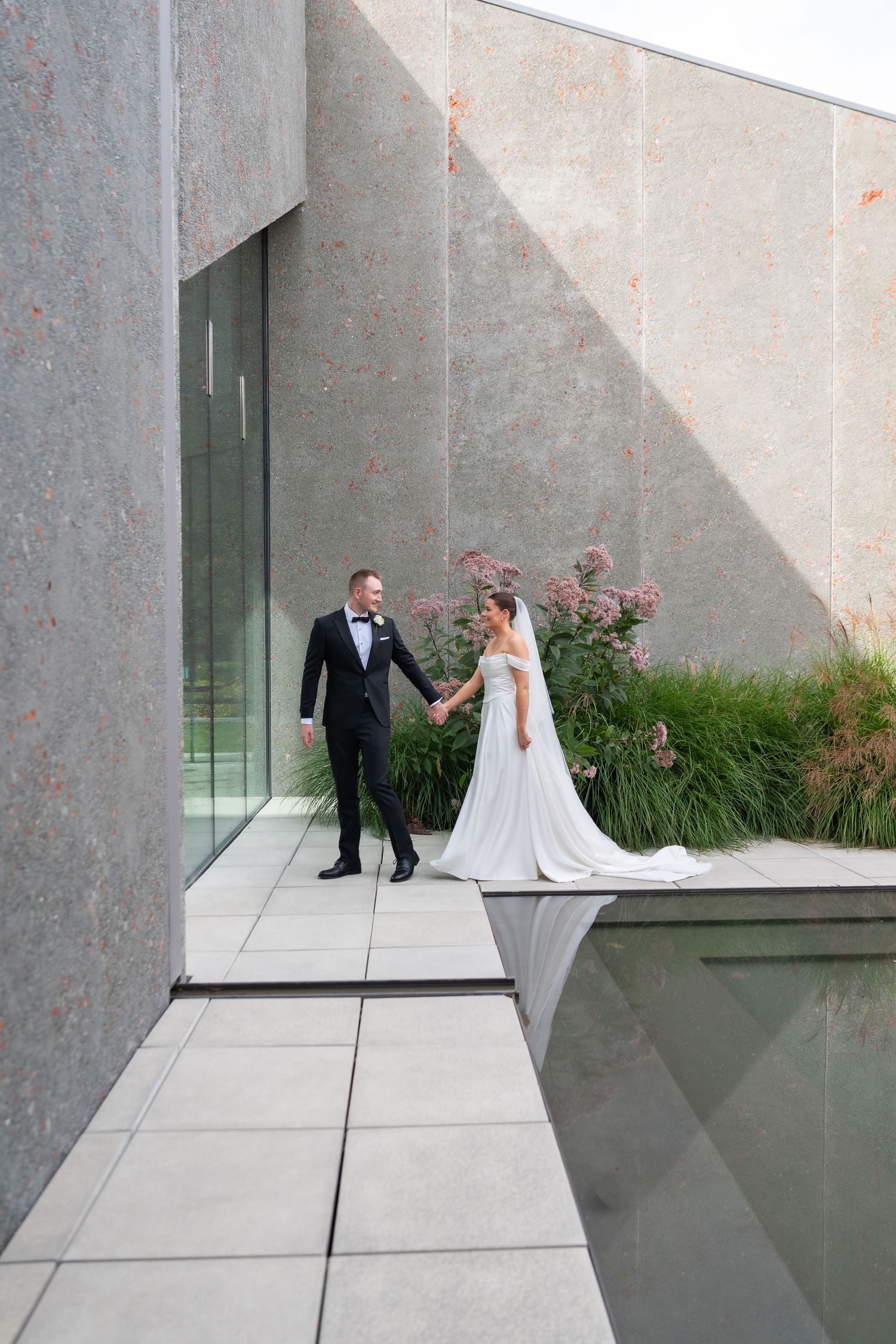 Bride and groom holding hands walking along modern stone building exterior with lush greenery on their wedding day.