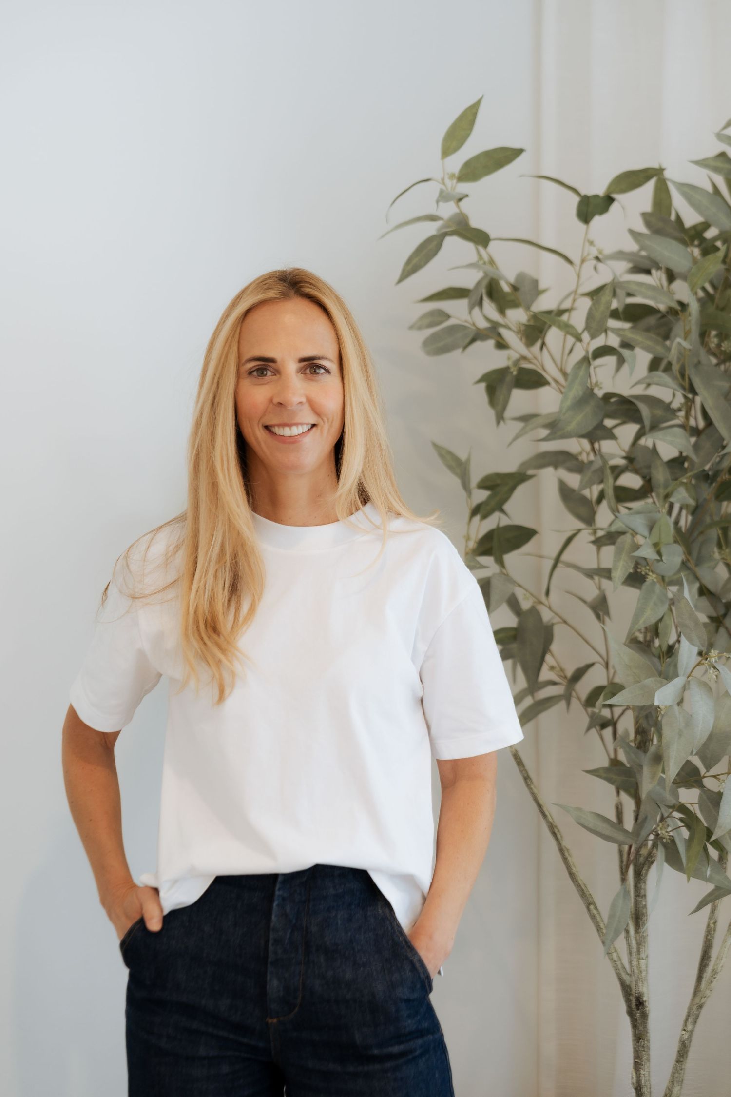 Smiling blonde woman in white t-shirt and jeans standing by a green plant against a light wall.