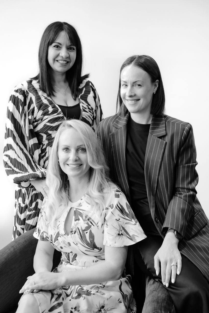 Black and white portrait of three smiling women, two standing and one seated, in stylish outfits against a light background.