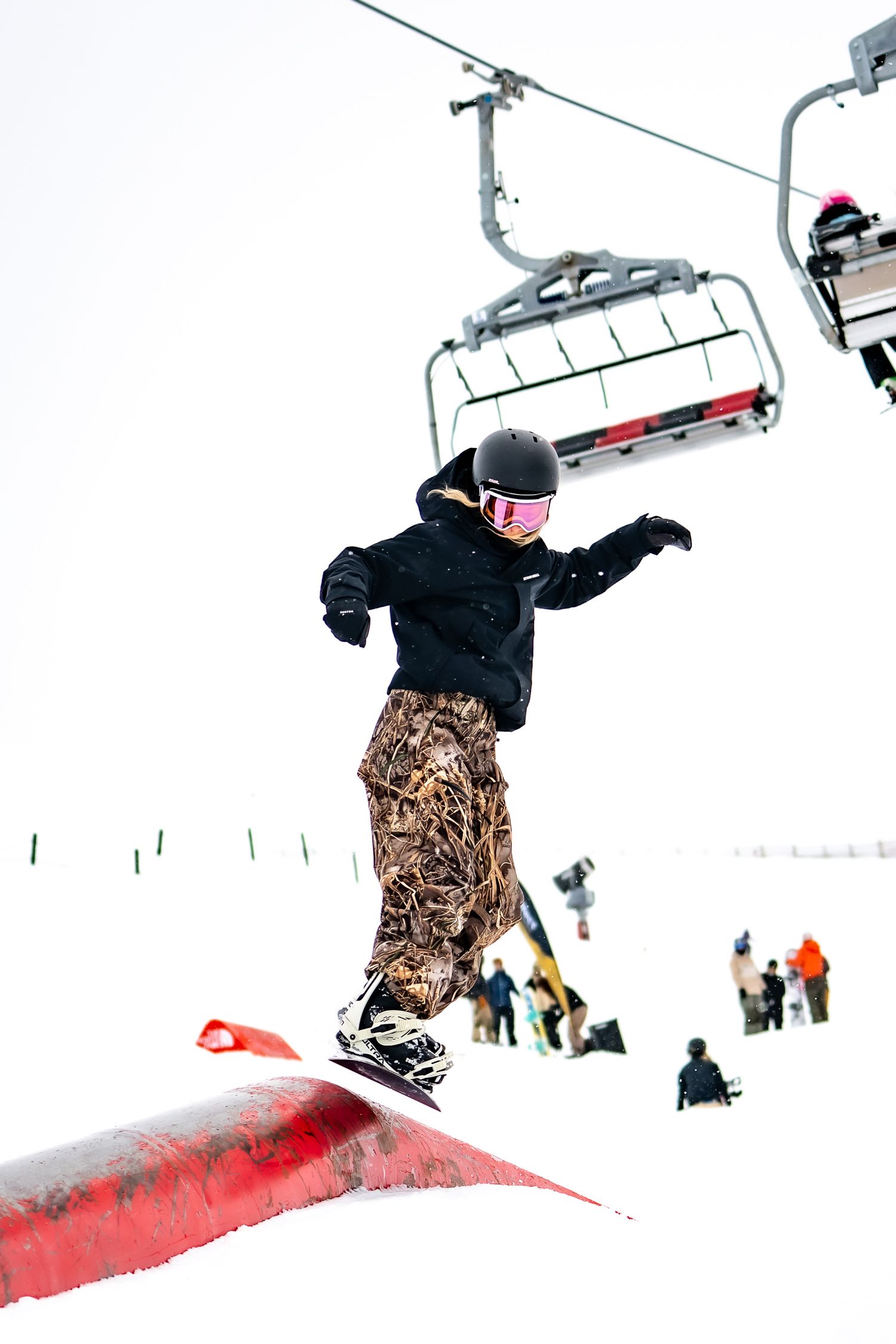 Female snowboarder performing trick jumps over red rail feature at ski resort with chairlift in background at Cardrona Alpine Report.