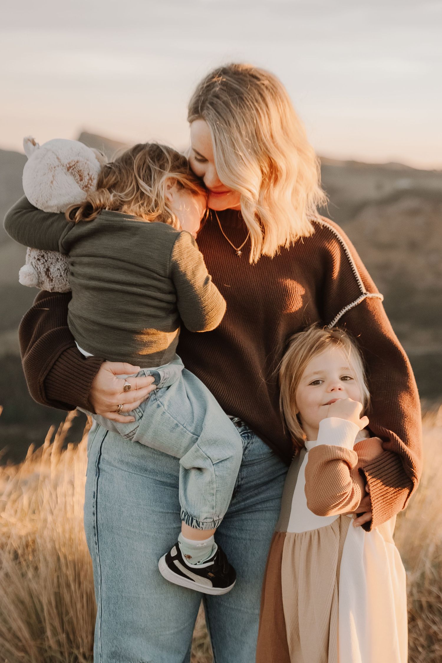 A blonde mother holds a toddler while a young girl in a cream dress stands beside her in a golden field at sunset.