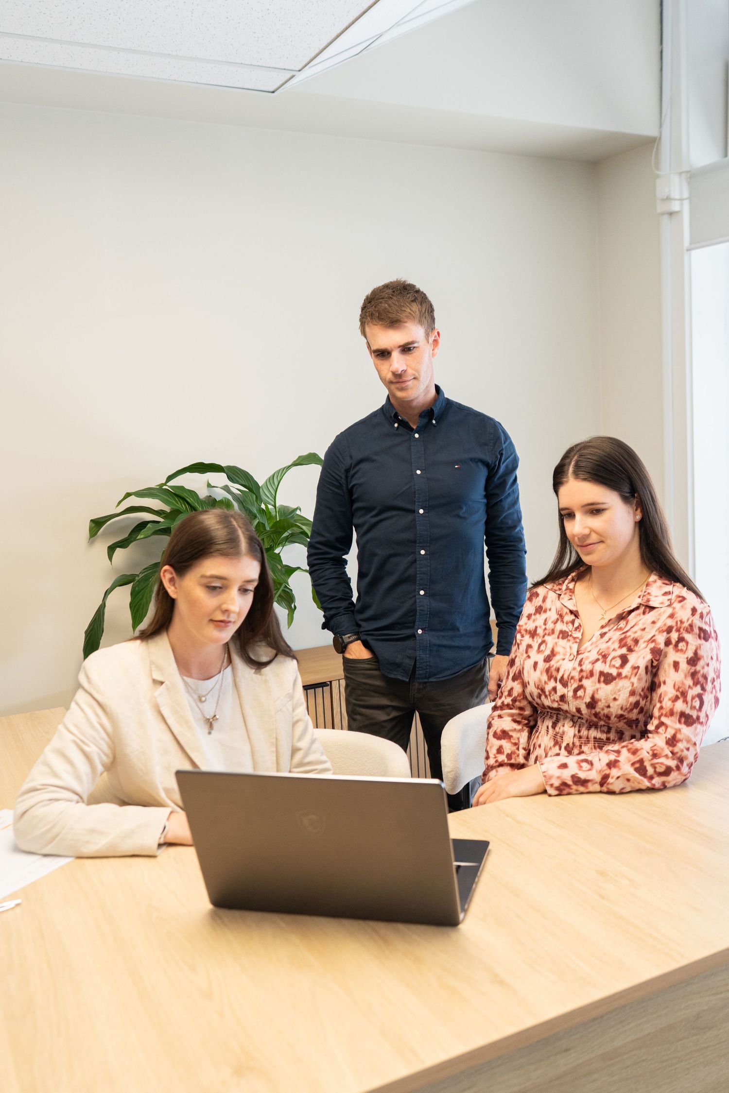 Three young professionals collaborating around a laptop at a wooden desk in a modern office setting.