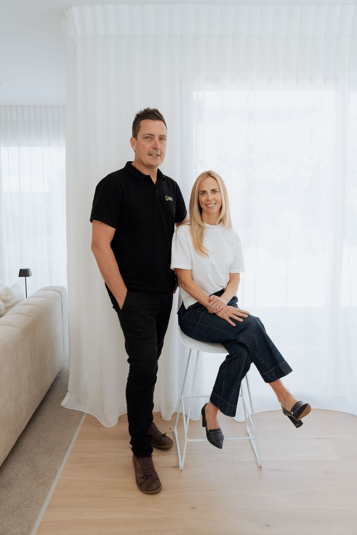 Man in black polo and woman in white t-shirt posing together in a bright modern living room.