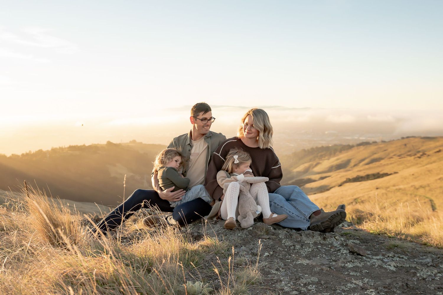 Family of four with a dog sitting on a sunny hillside at golden hour with scenic valley views.
