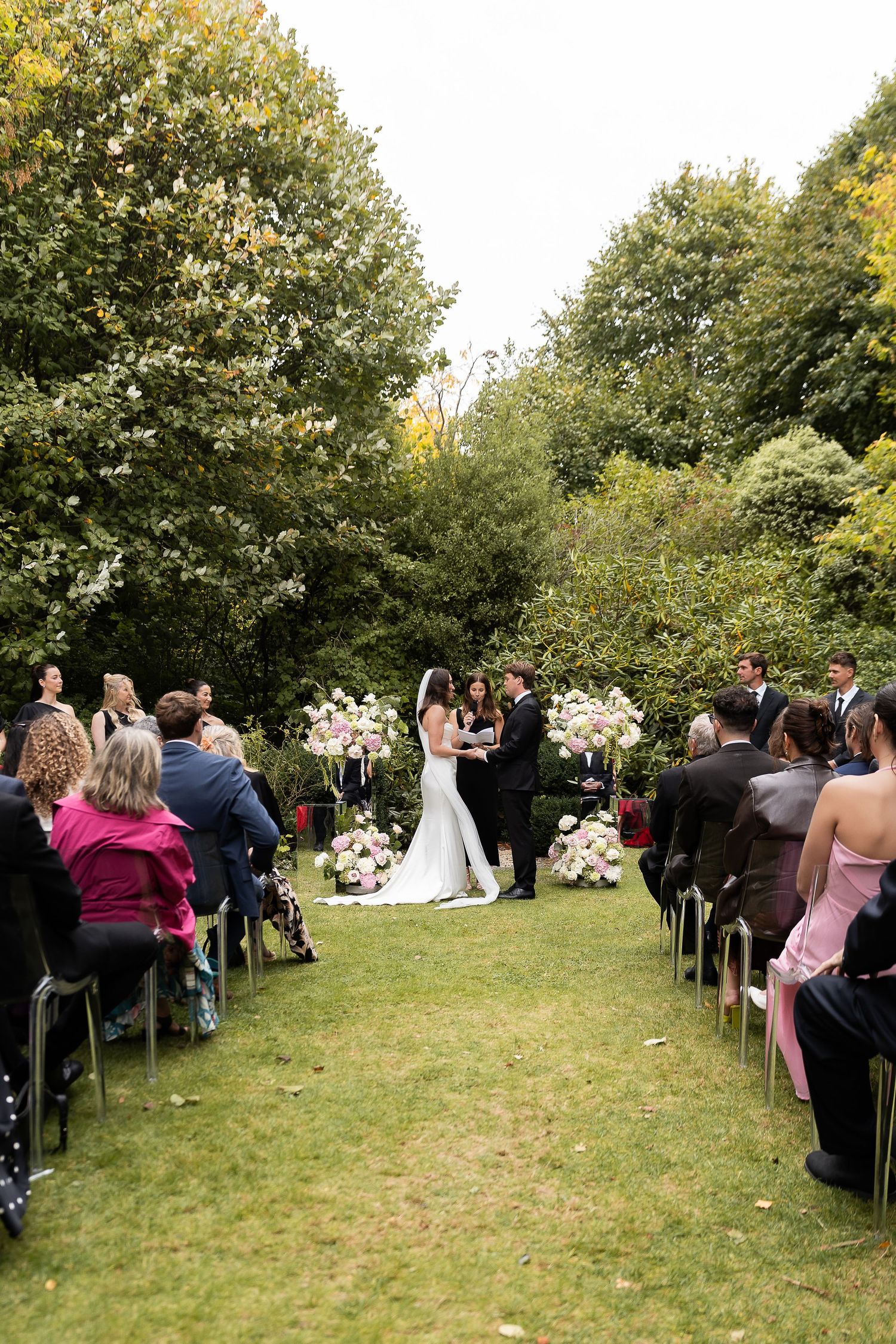 Outdoor wedding ceremony on a green lawn with guests seated in rows, couple at the altar surrounded by lush trees and floral arrangements.