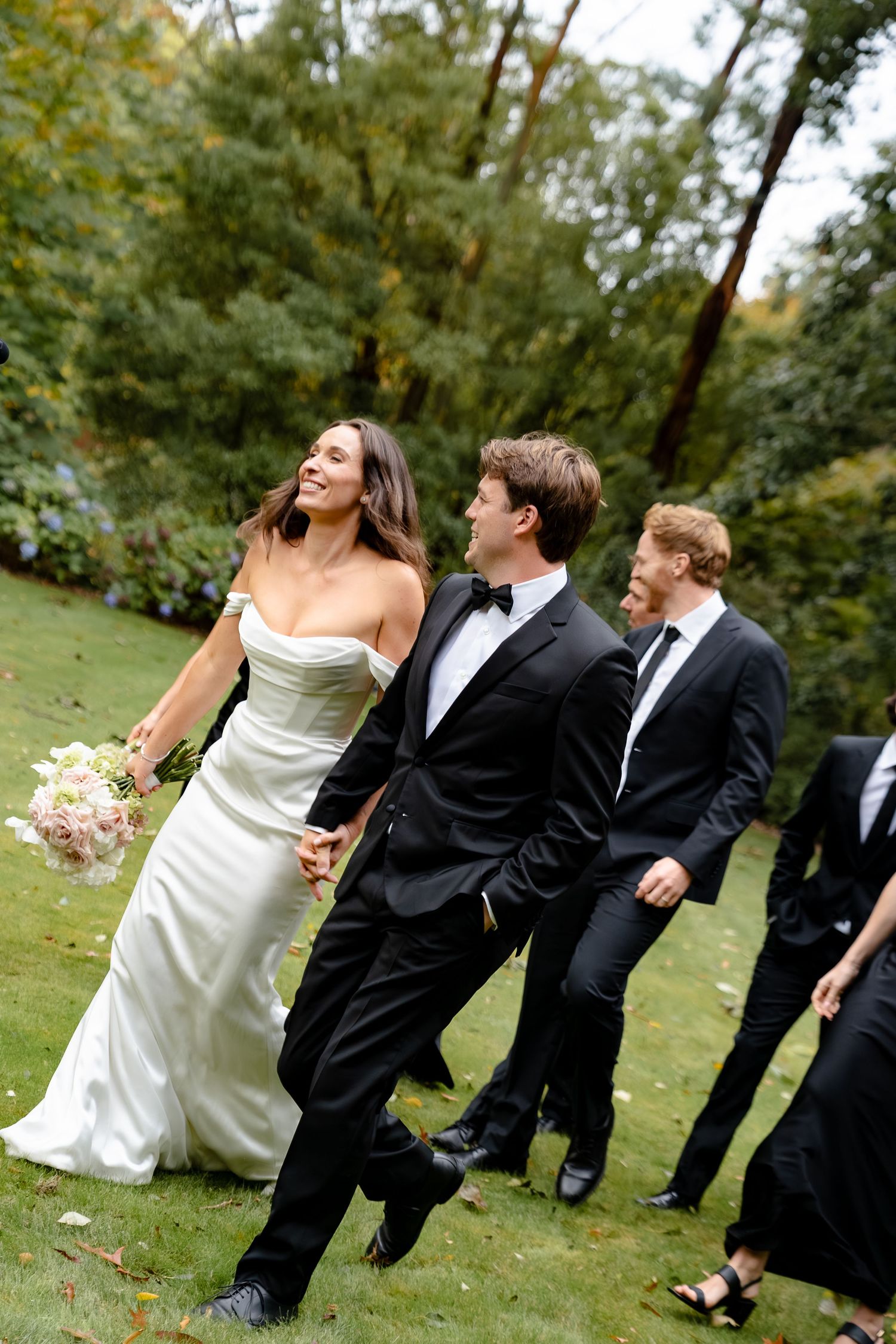 Bride in white gown holding bouquet walks with groom in black tuxedo and groomsmen on green lawn.