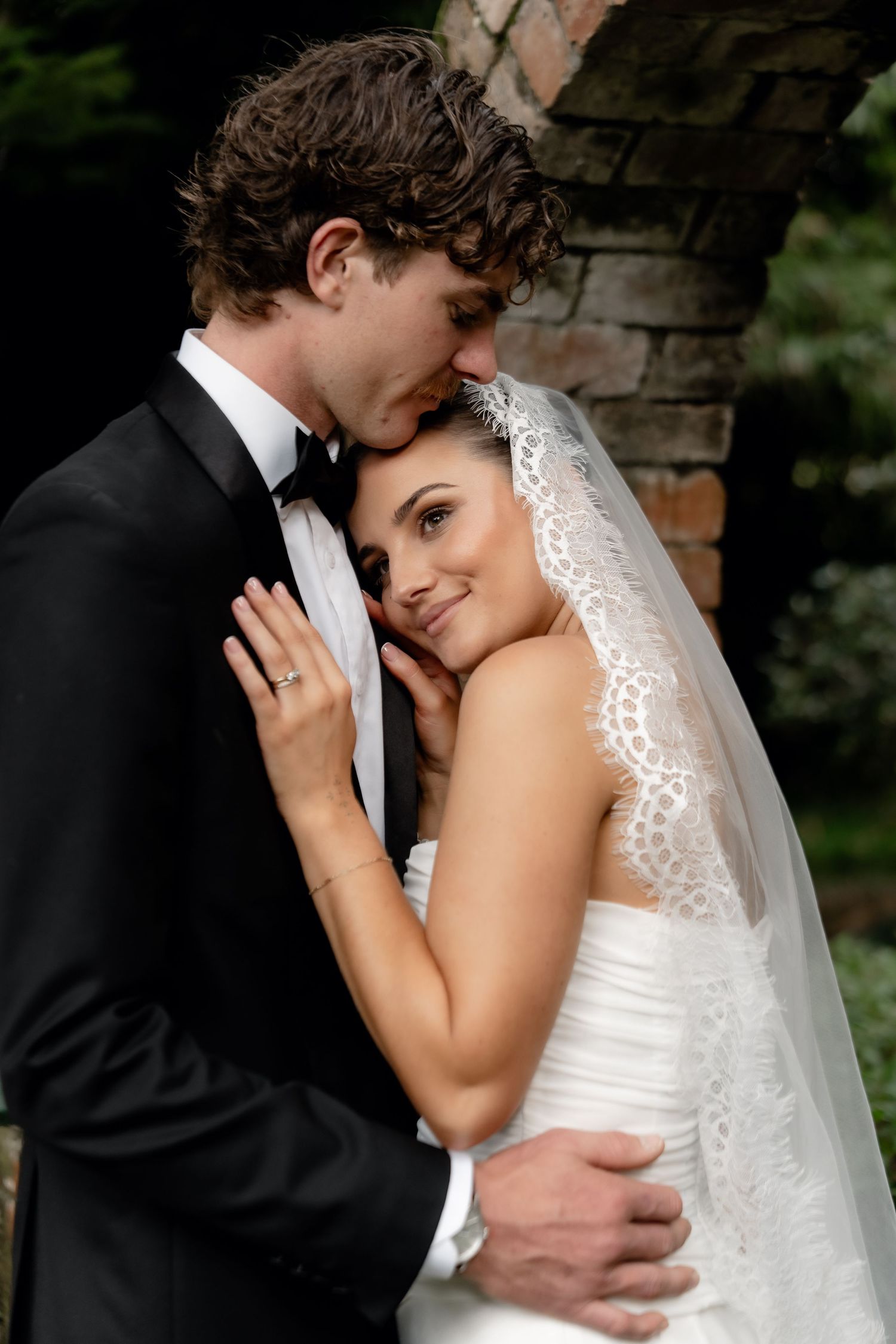 A bride in a white lace veil and gown embraces a groom in a black tuxedo outdoors near a brick wall.