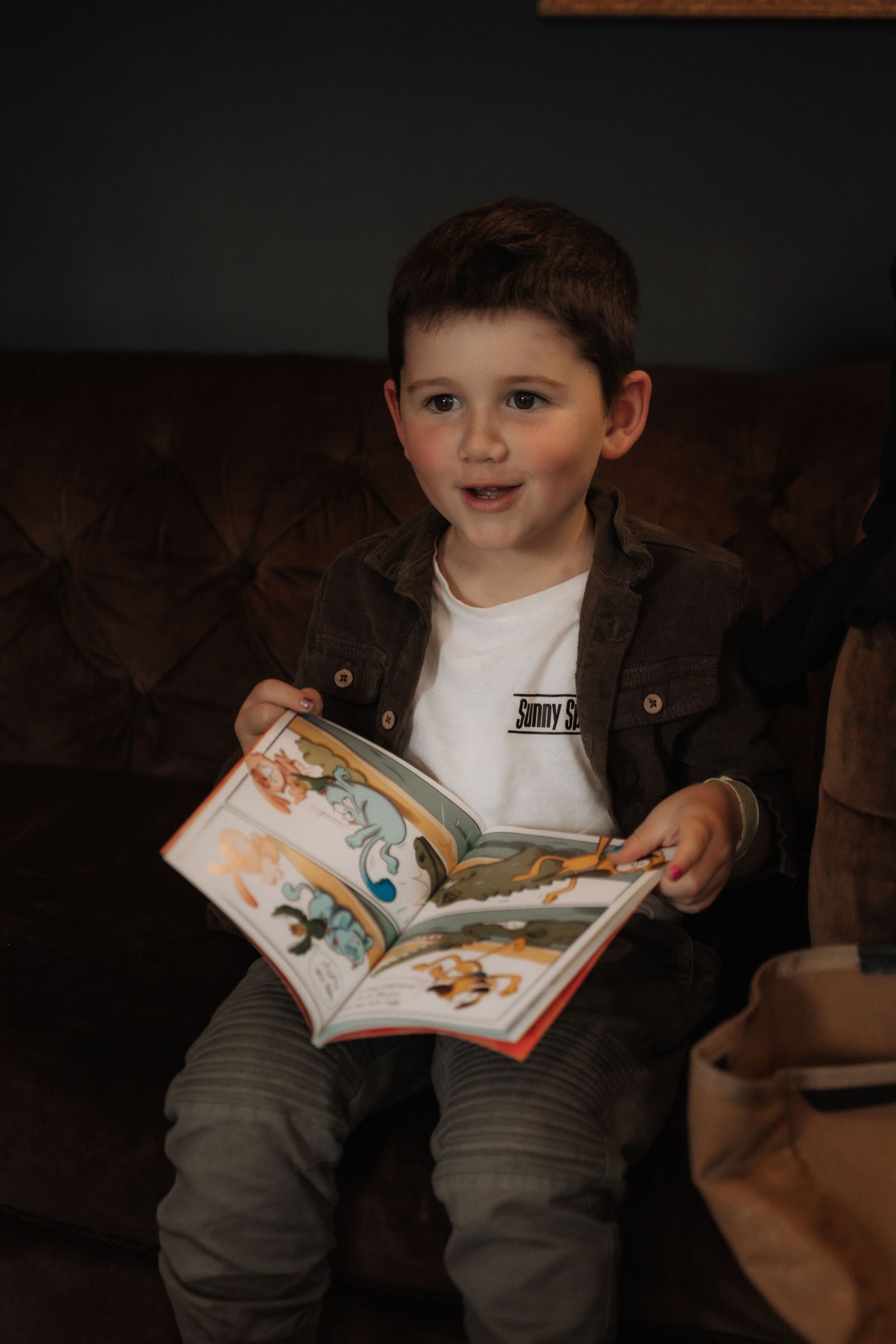 Young boy sitting on a sofa holding an open illustrated children's book, wearing a dark jacket and white shirt.