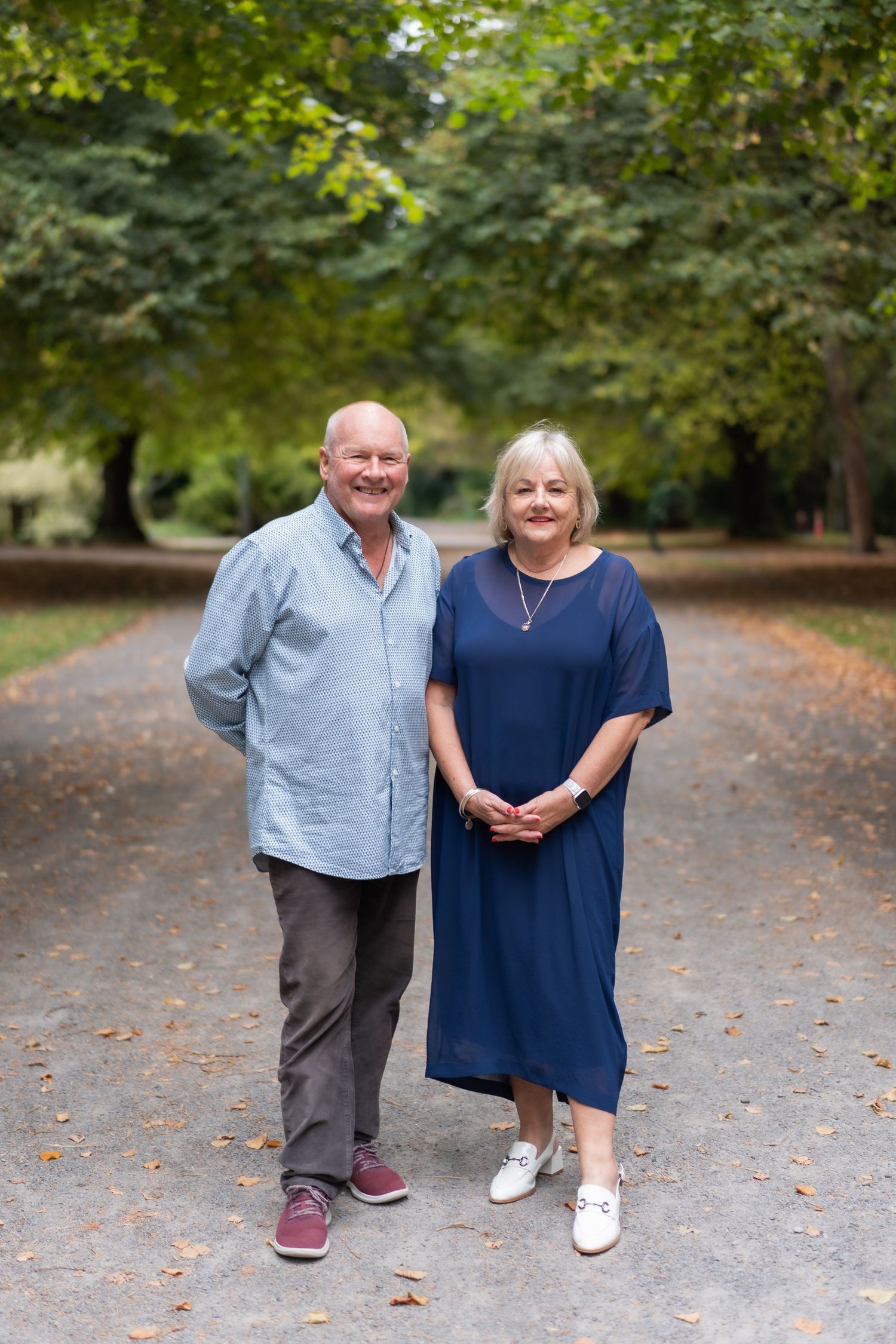 Smiling senior couple standing together on a tree-lined park path on a sunny day.