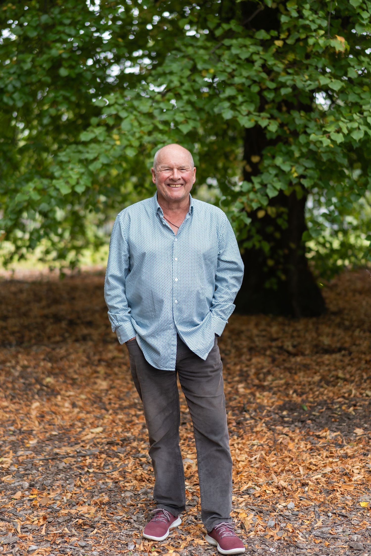 Smiling senior man in blue shirt and gray pants standing outdoors under a large green tree on a bed of fallen leaves.