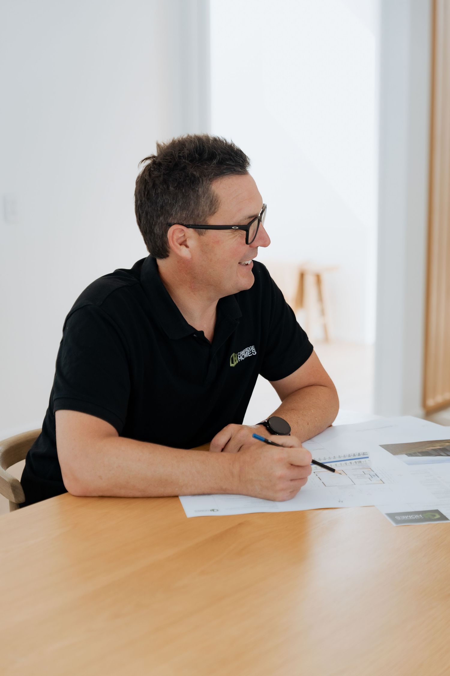 Man in black polo shirt with glasses sitting at wooden table reviewing documents with a pen in a bright office.