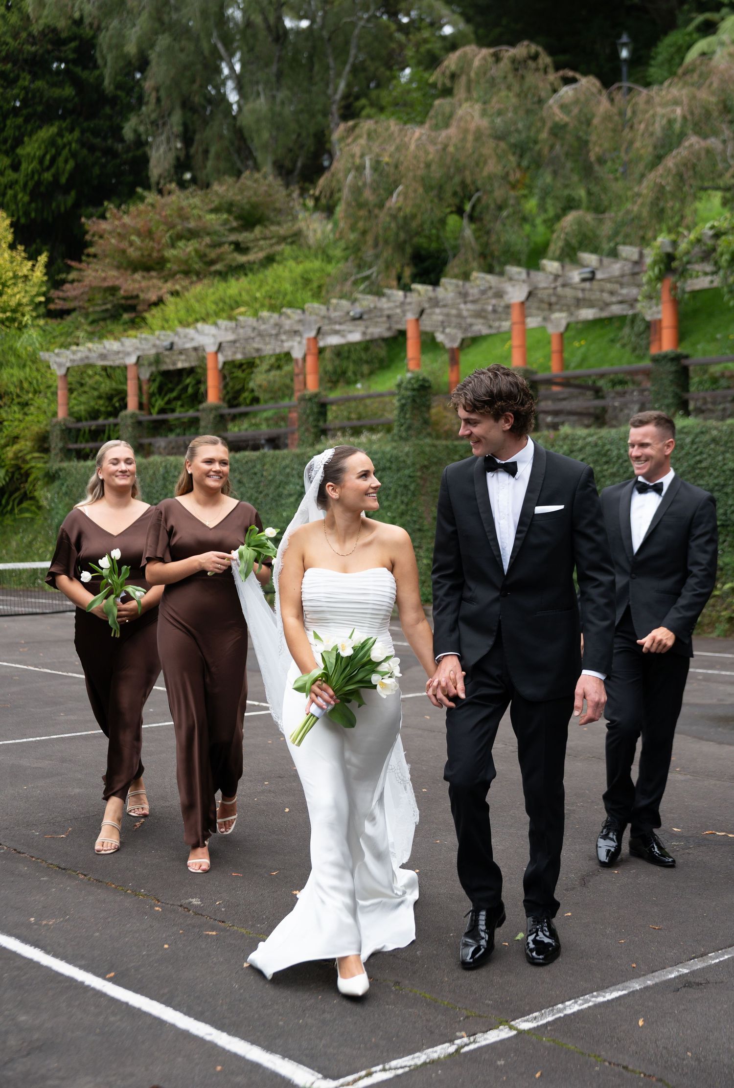 Bride and groom walk hand in hand with bridesmaids in brown dresses and groomsmen in black tuxedos outdoors.