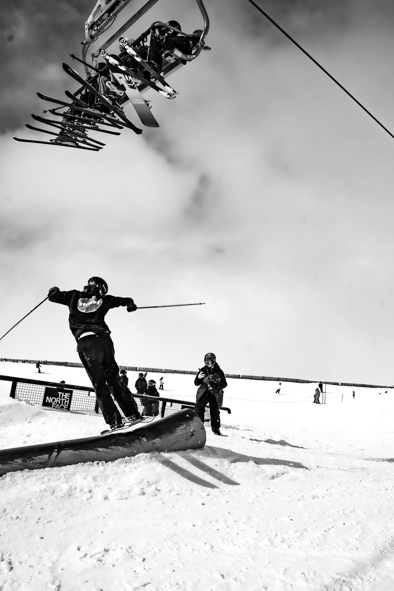 A skier performs a rail slide trick at a snow park while a chairlift carries riders overhead in black and white photography at Cardrona.