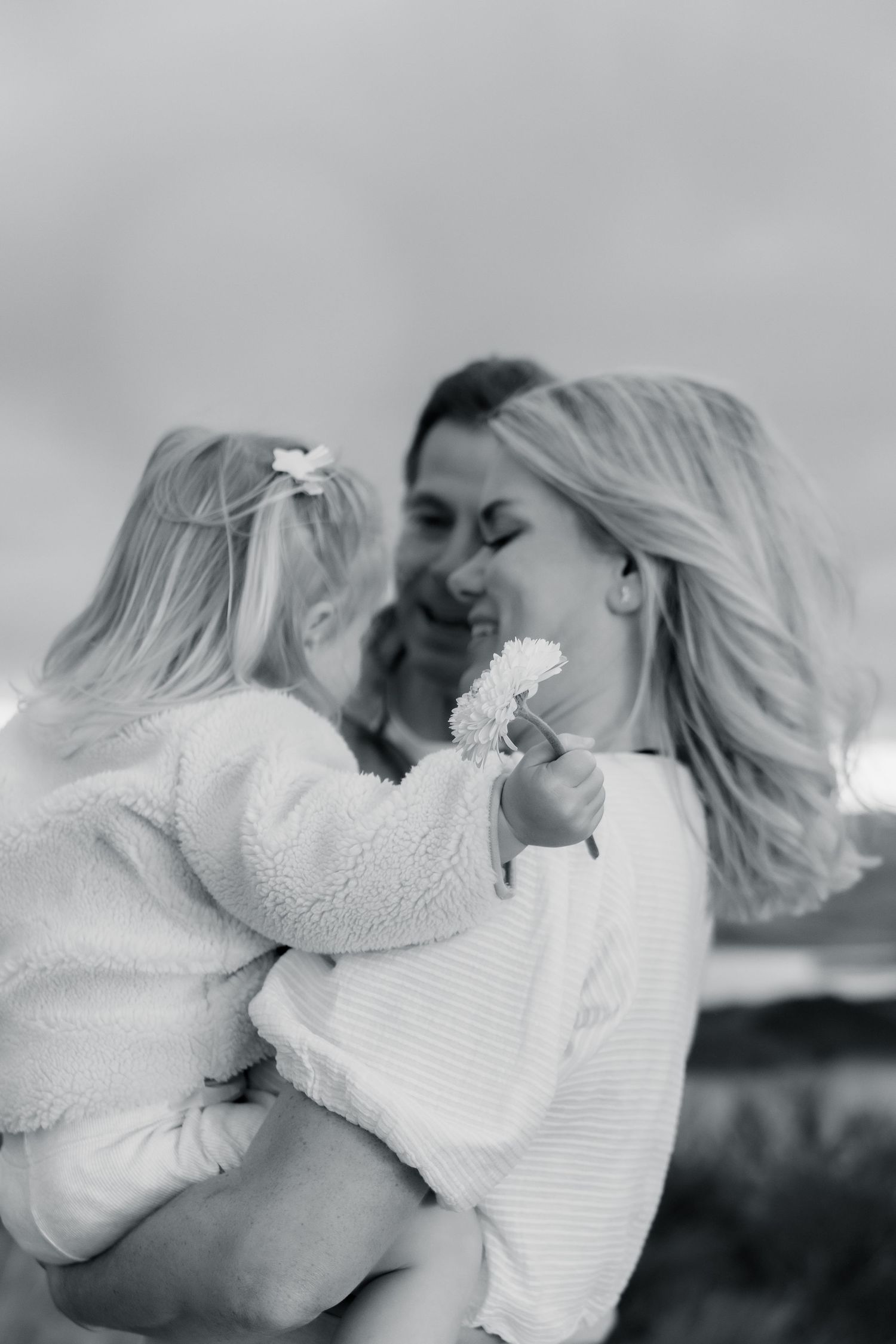 Black and white photo of a smiling mother holding her young daughter who offers her a daisy flower outdoors.