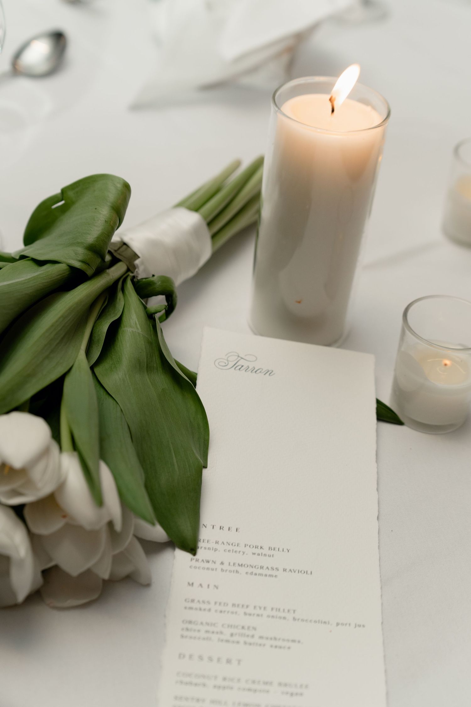 Elegant wedding table setting with white tulips, lit candle, and a printed menu card on white linen.