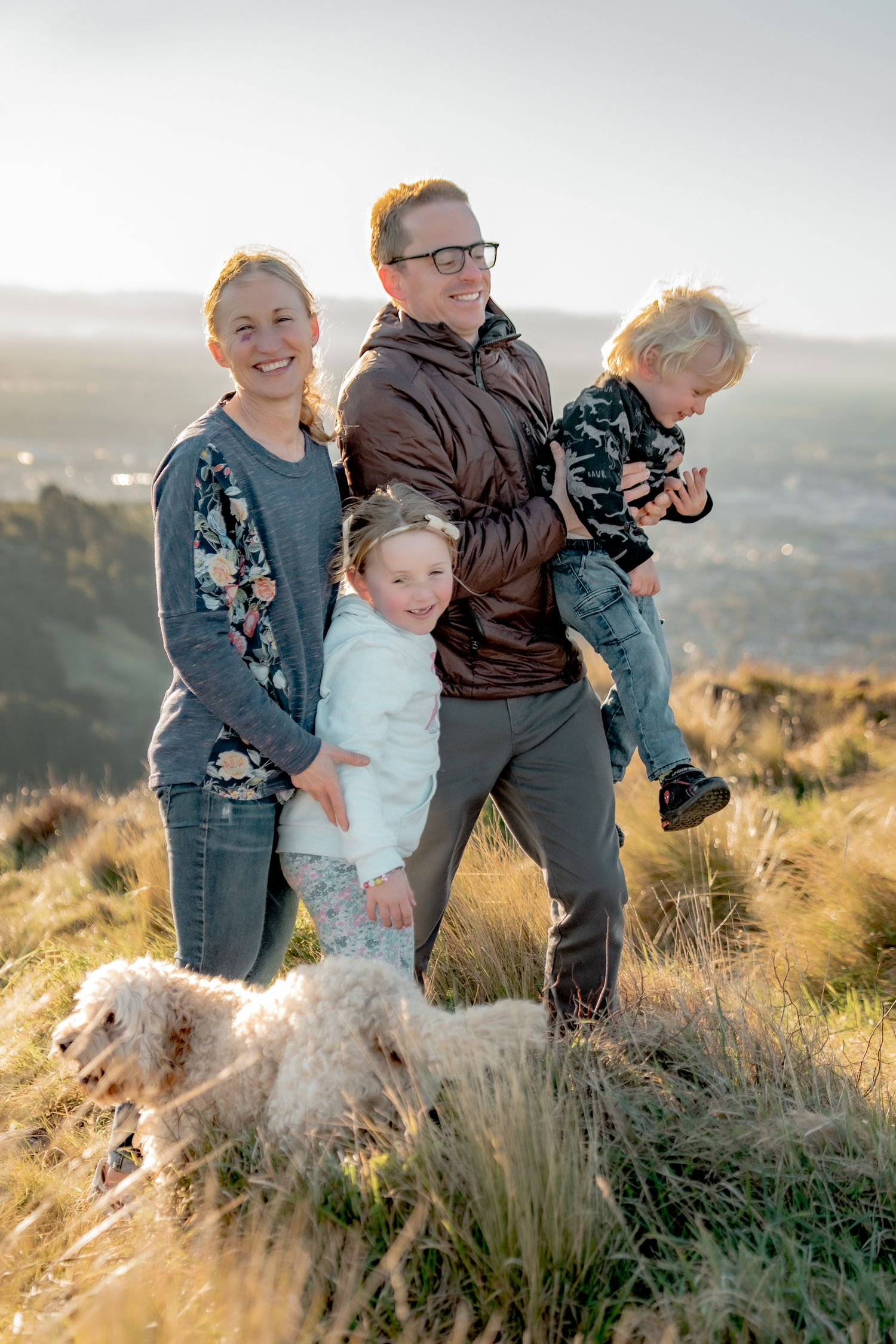A family and their fluffy white dog enjoy a golden sunset moment together on a grassy hilltop overlook.