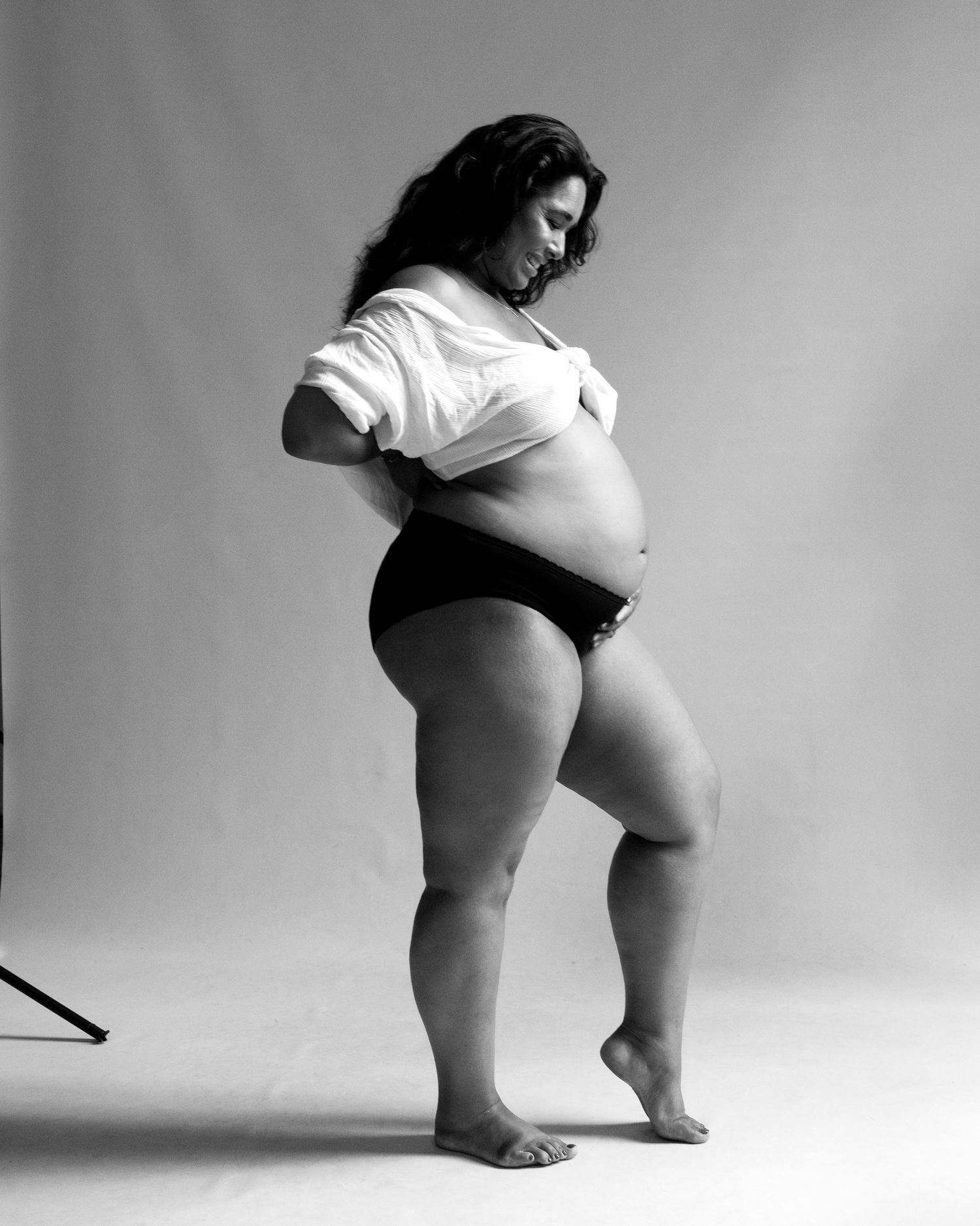 Black and white artistic portrait of a pregnant figure in cropped white top and dark underwear against neutral backdrop.