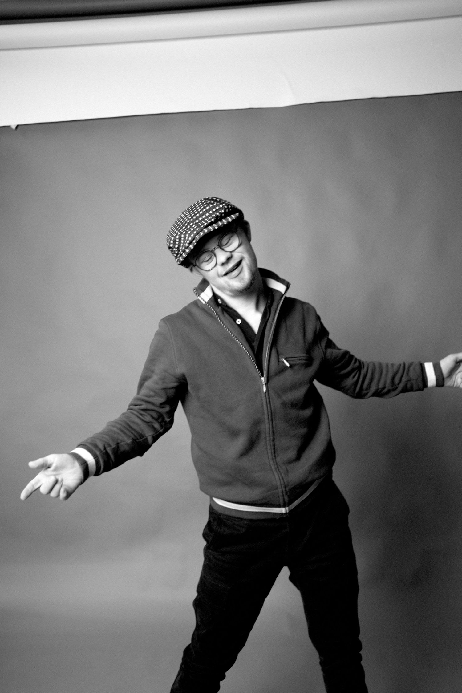 Black and white portrait of a person in a flat cap and zip-up sweater striking a playful pose against a dark background.