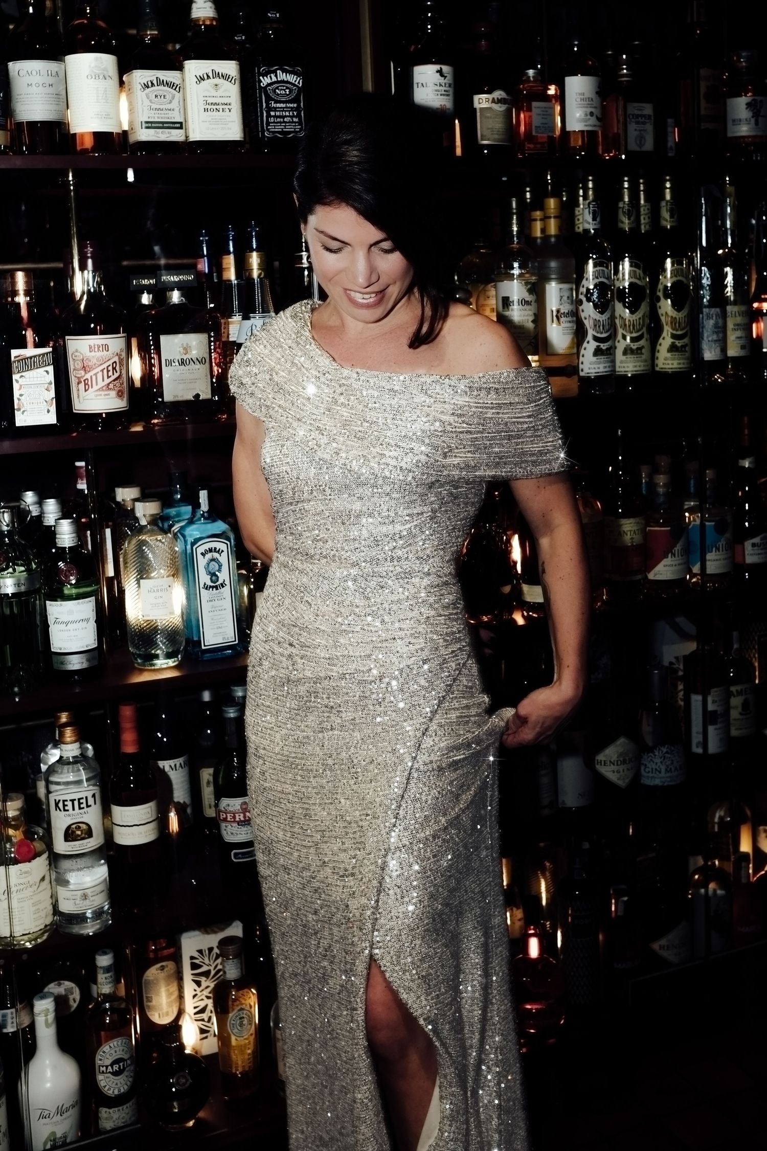 A person in a sparkly silver dress poses at a bar with shelves of liquor bottles in the background.
