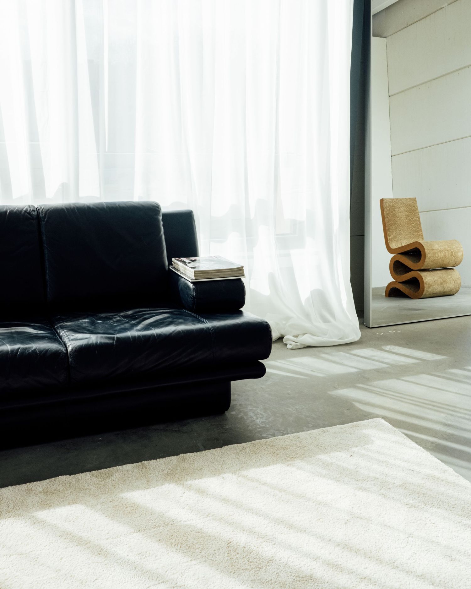 Minimalist living room with black leather sofa, sheer white curtains, a wiggle chair, and soft natural light on a neutral rug.