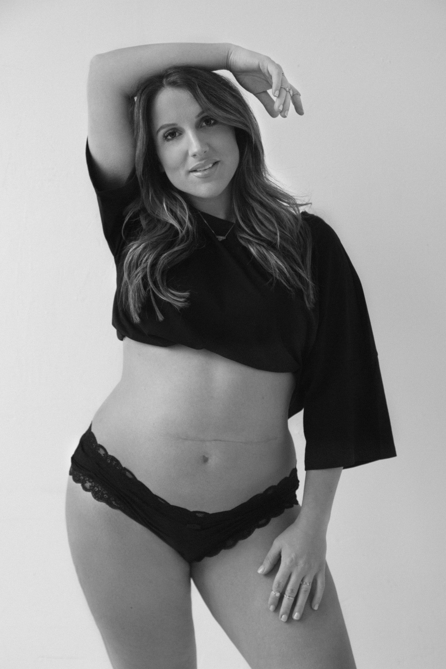 Black and white boudoir photo showing a person in a cropped top and lace underwear posing against a white background.