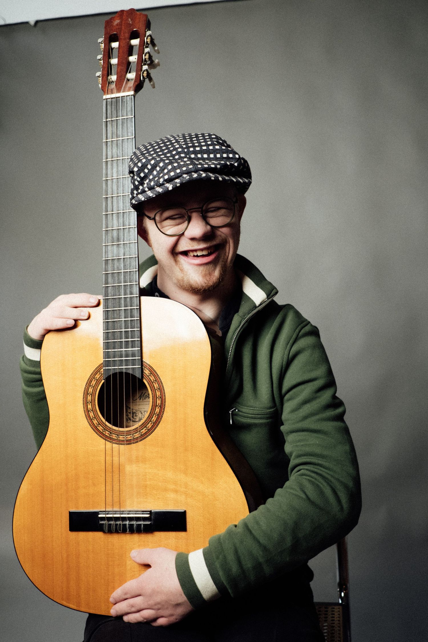 Smiling young man holding an acoustic guitar, wearing glasses and a flat cap, against a gray background.