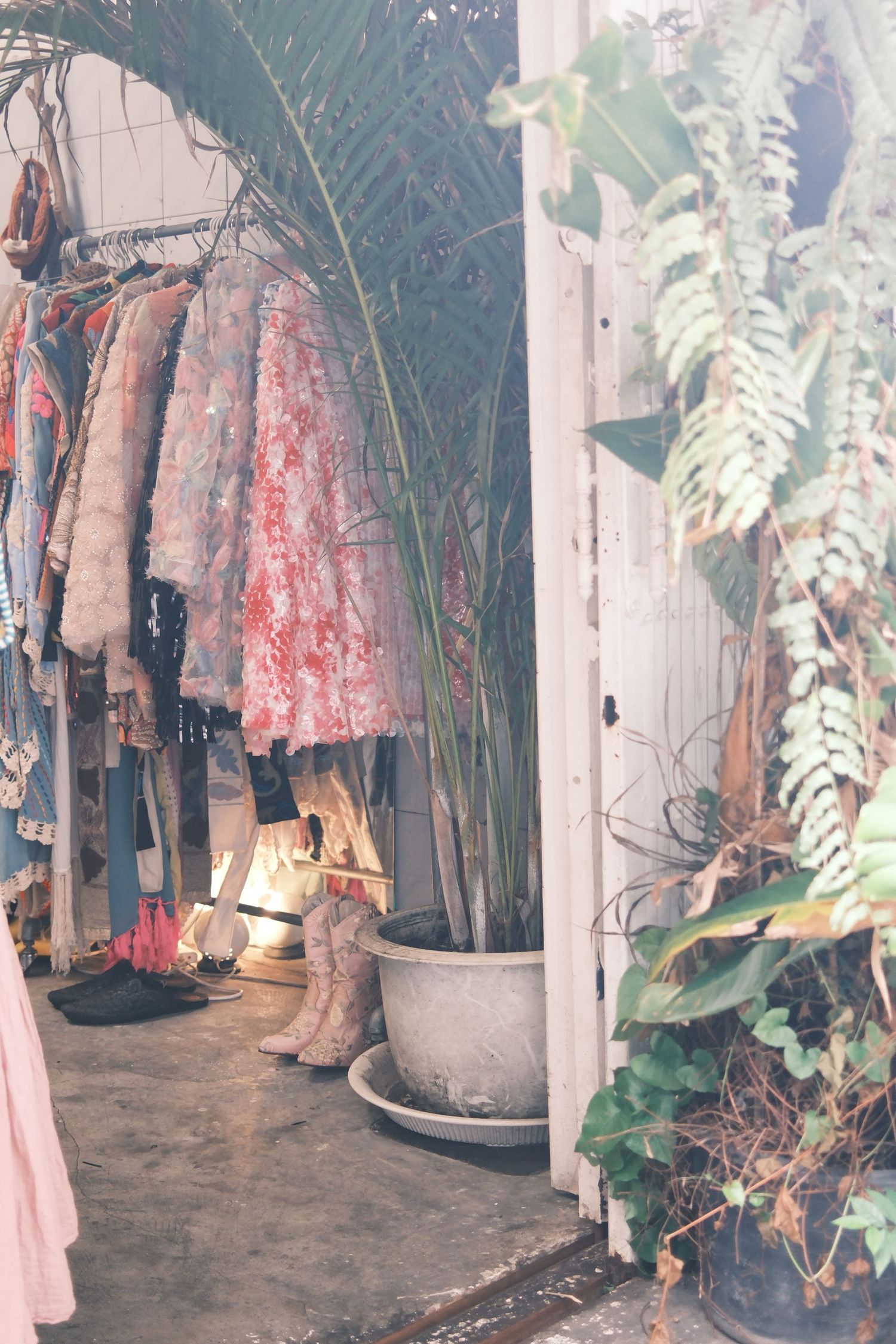 Boho boutique with floral dresses on rack, cowboy boots, potted palm tree, and lush green plants by white door.