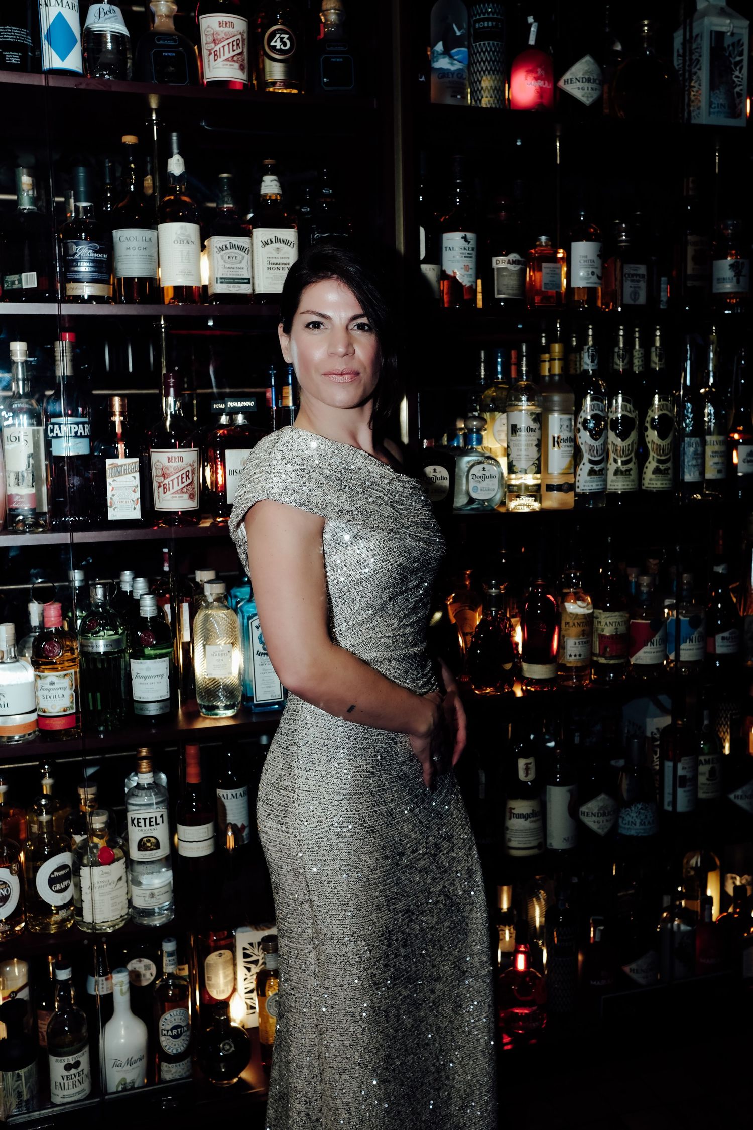 A person in a sparkly silver evening gown poses in front of a well-stocked bar with illuminated shelves of liquor bottles.