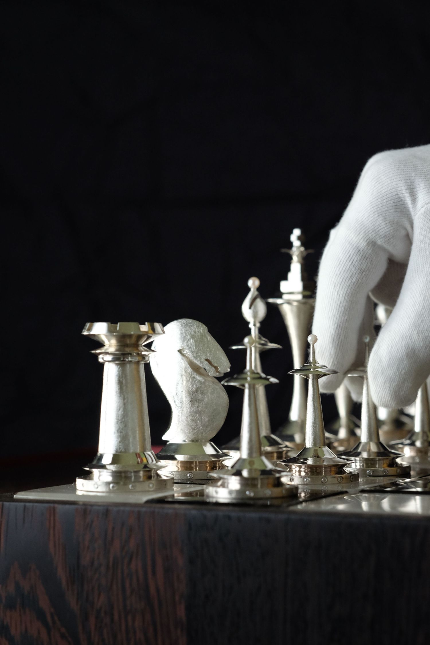 White chess pieces arranged on a dark wooden surface with a gloved hand moving pieces in dramatic lighting.