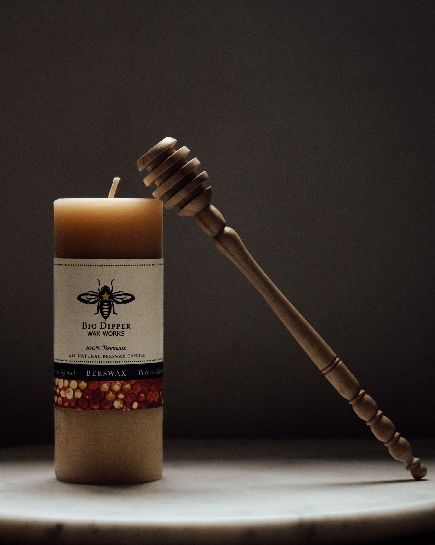 A wooden honey dipper rests beside a beige candle with a bee logo and red beaded decorative elements in moody lighting.