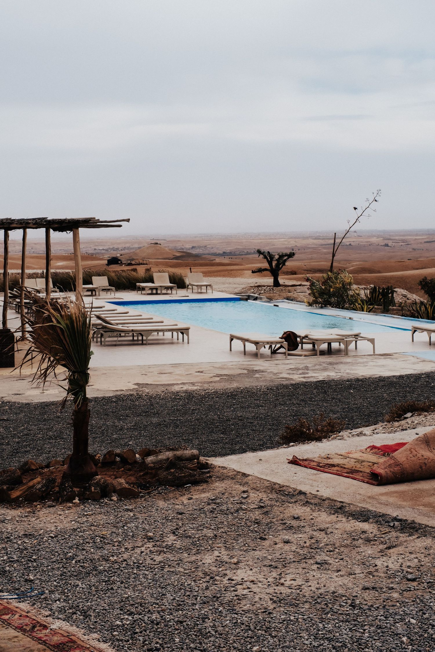Luxurious desert resort swimming pool with lounge chairs overlooking vast Moroccan sand dunes under cloudy skies.