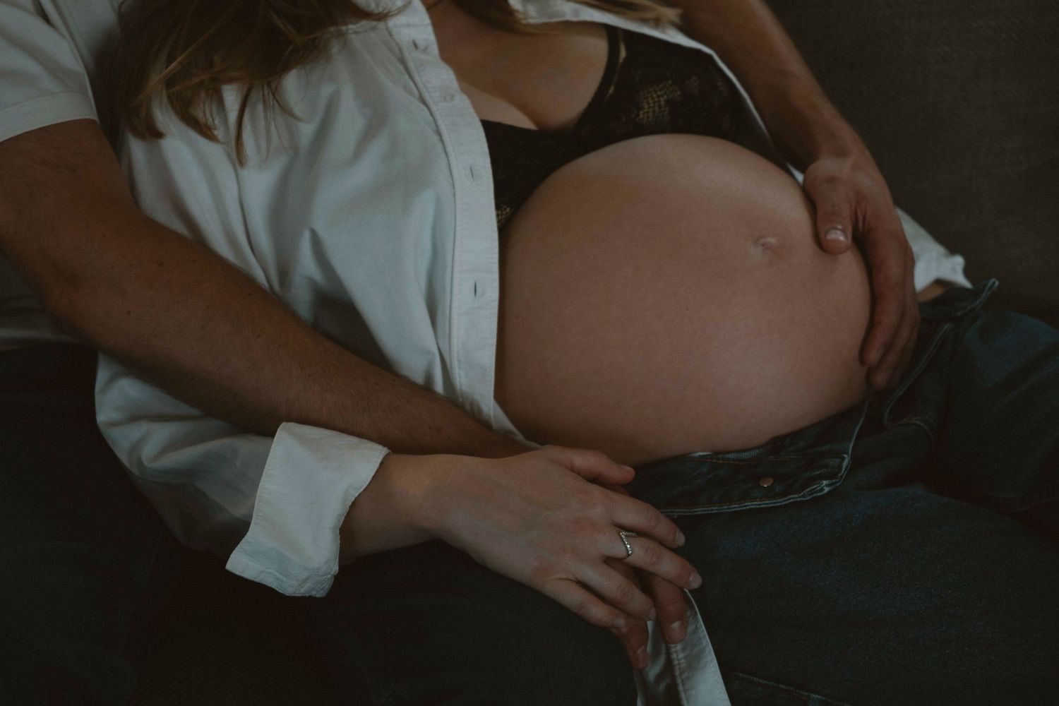 A pregnant person's belly is shown while resting on a dark couch, wearing a white shirt and dark pants.
