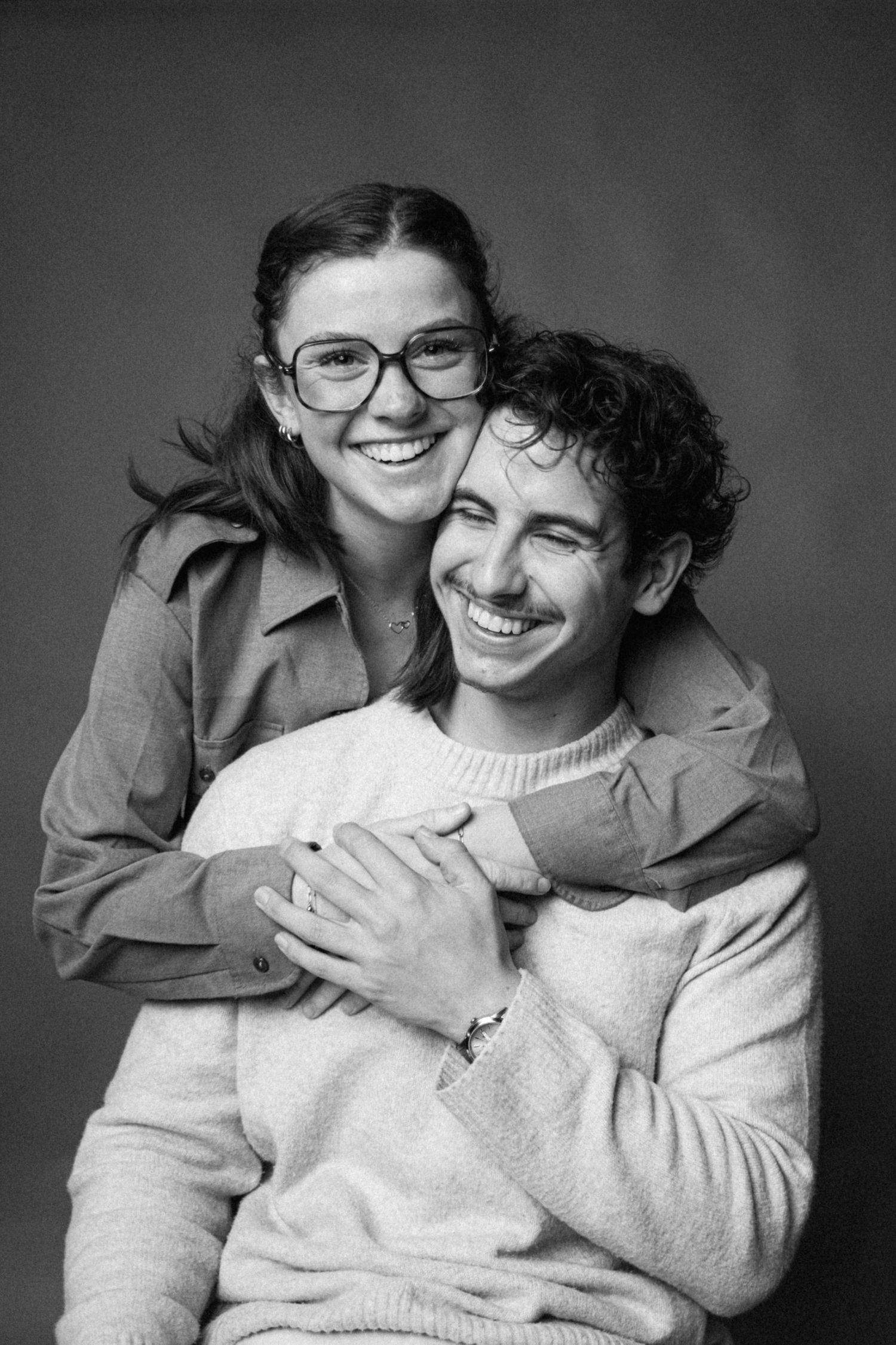 Black and white portrait of a couple embracing and smiling, with one wearing glasses and a jacket, the other in a sweater.