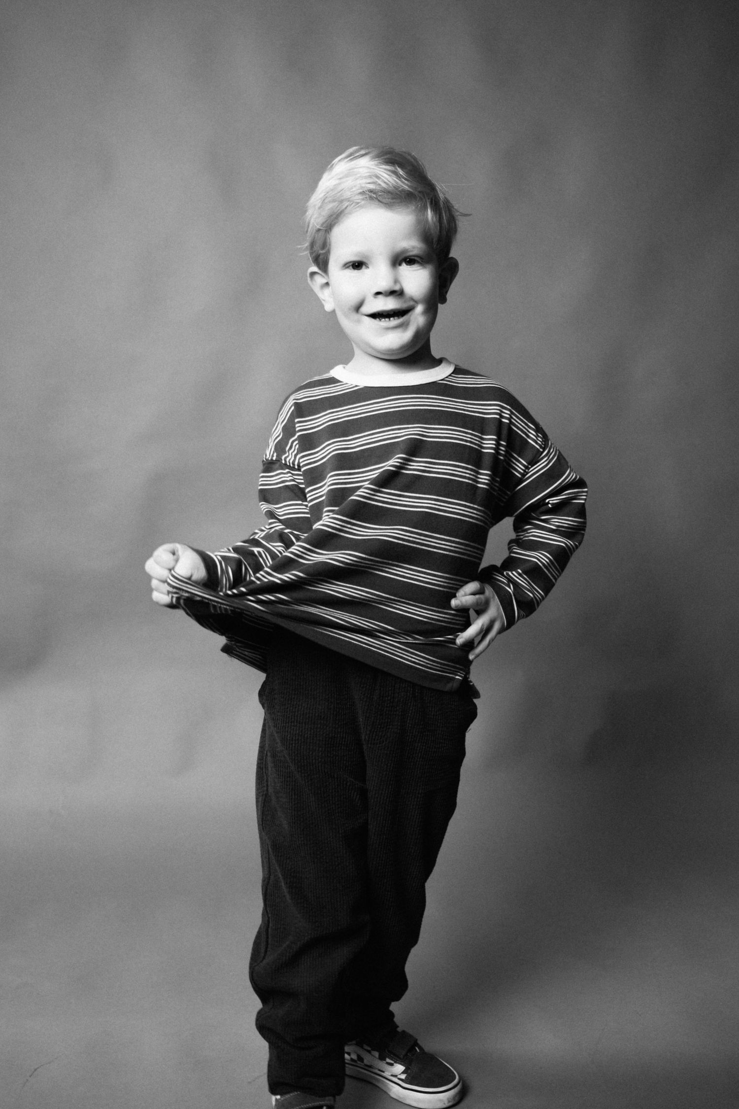 Black and white studio portrait of a young child in a striped shirt and dark pants posing with a happy expression.