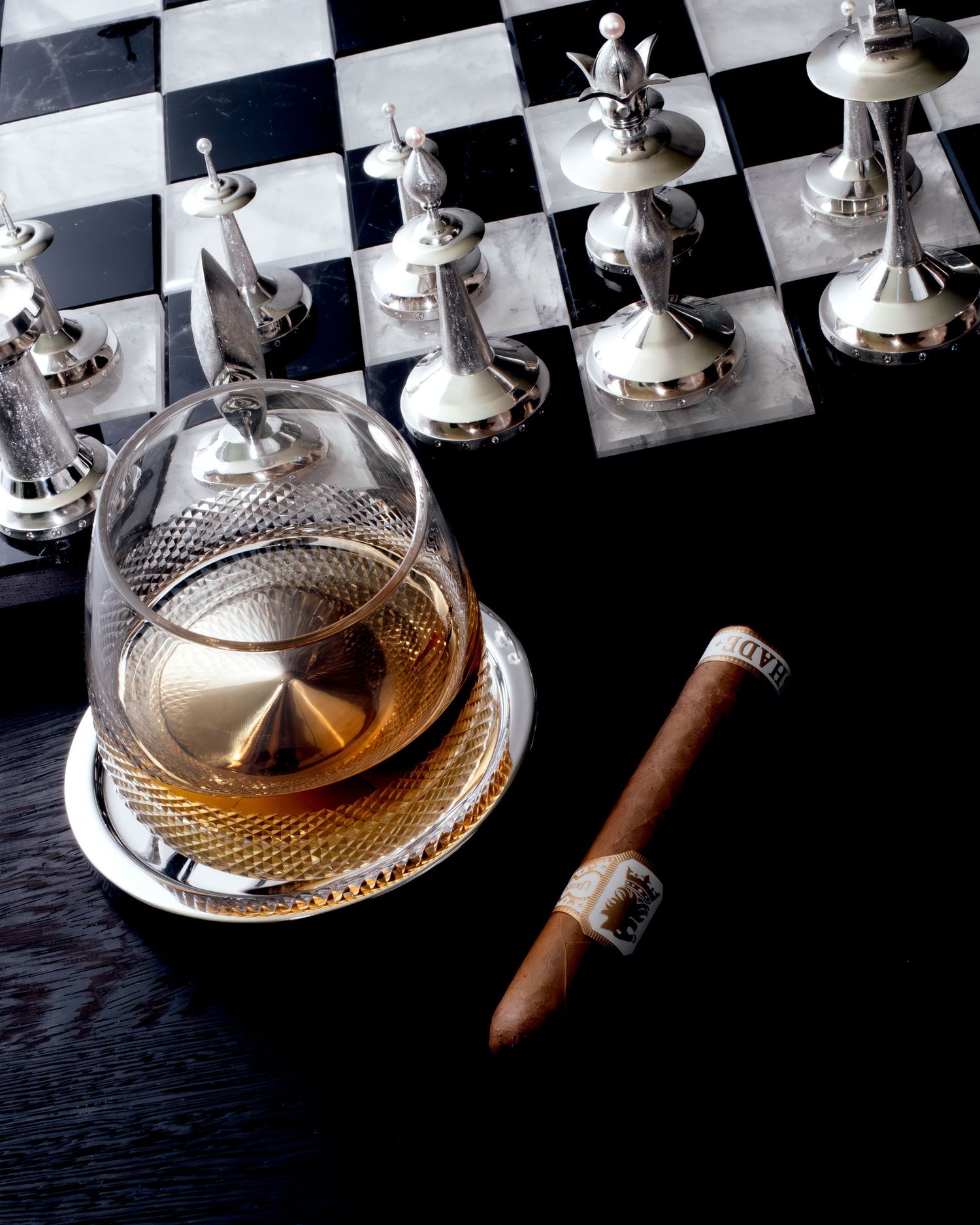 A silver tea service set with ornate detailing reflects light against a dark background with a cigar beside it.