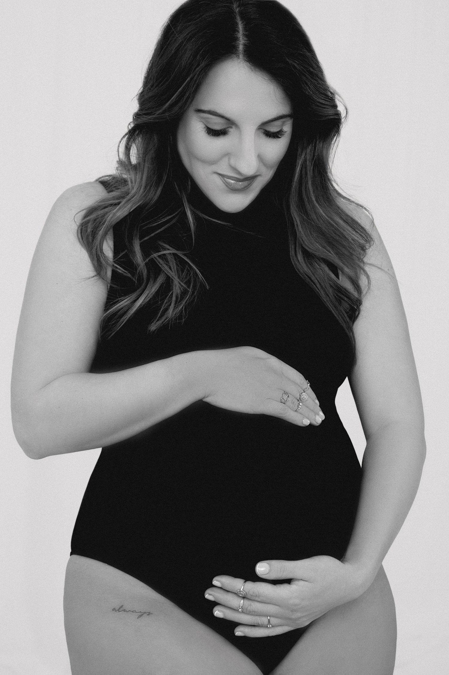 Black and white maternity photo of a woman in a dark bodysuit holding her pregnant belly with a gentle smile.