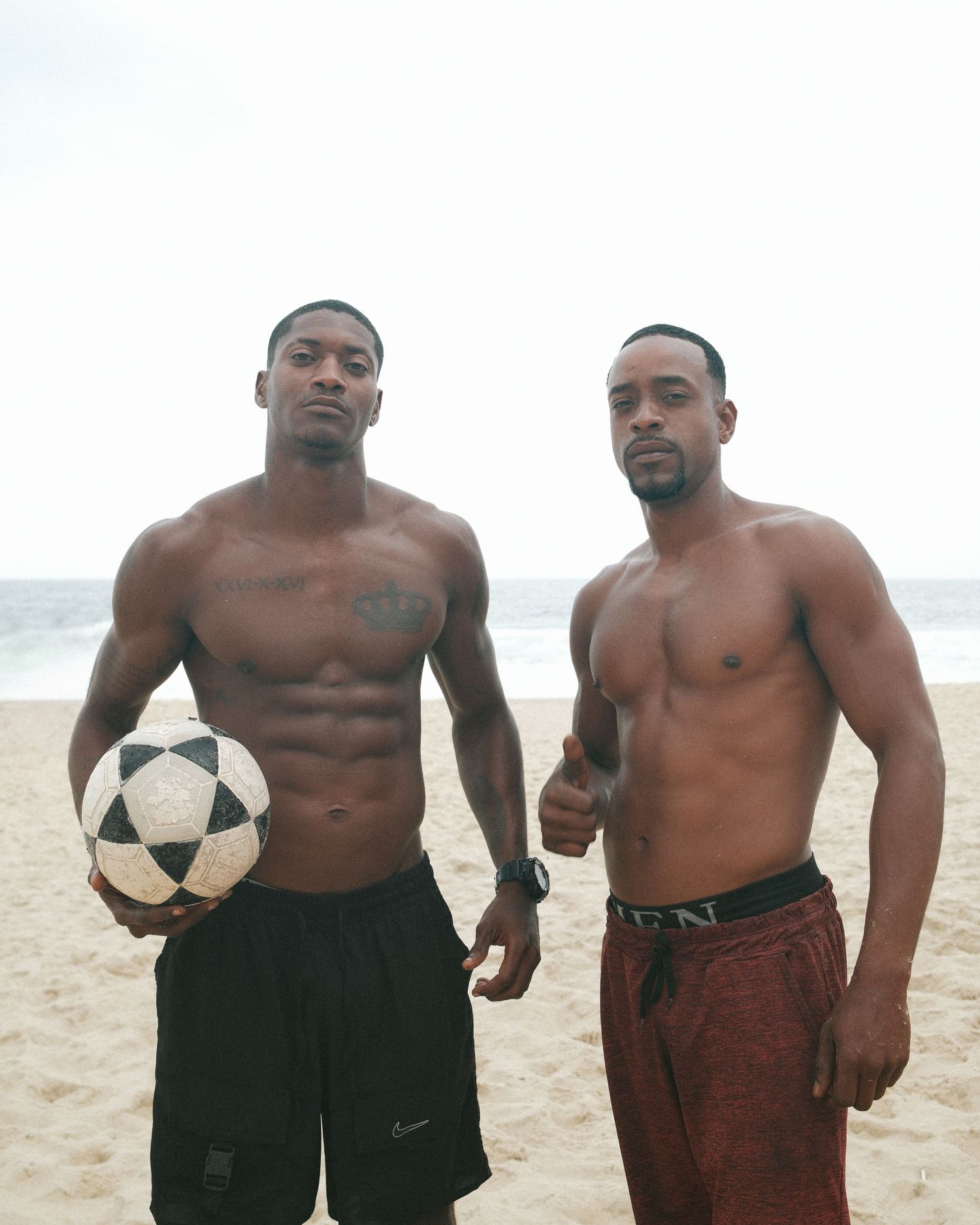 A group of athletic men holding soccer balls pose together on a sandy beach.