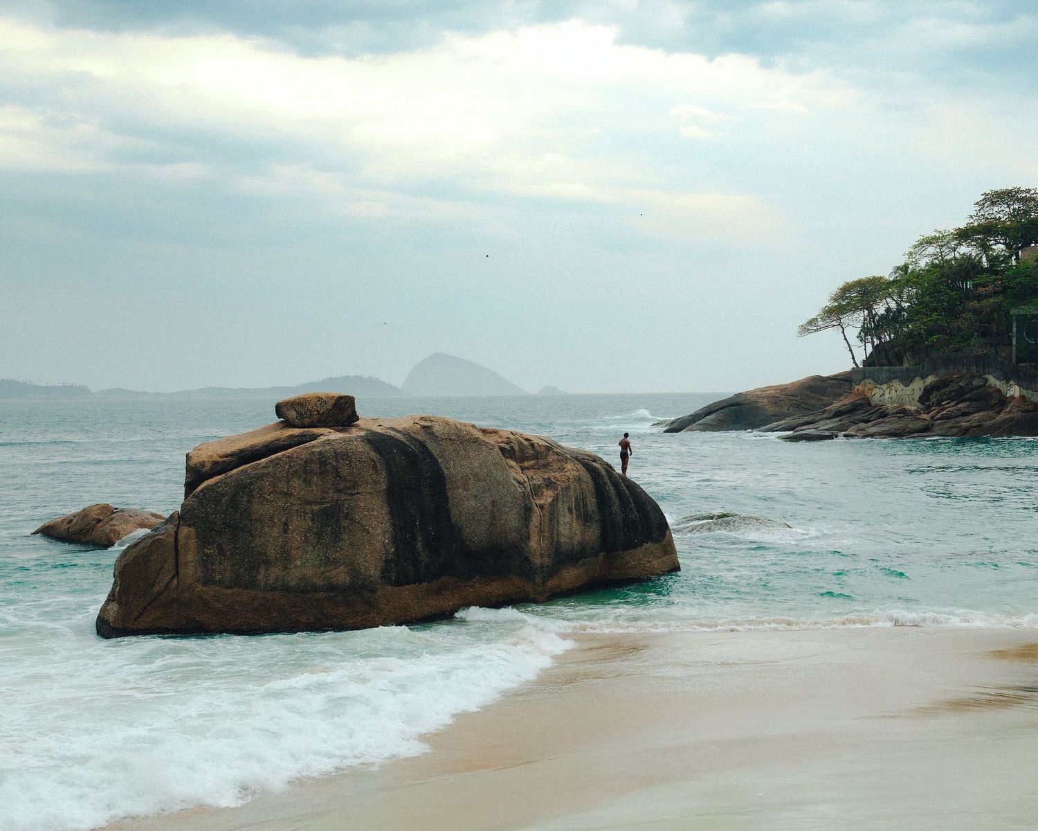 Dramatic panoramic view of a rocky beach coastline with large boulders and crashing waves against a cloudy sky, palmtrees and a man.