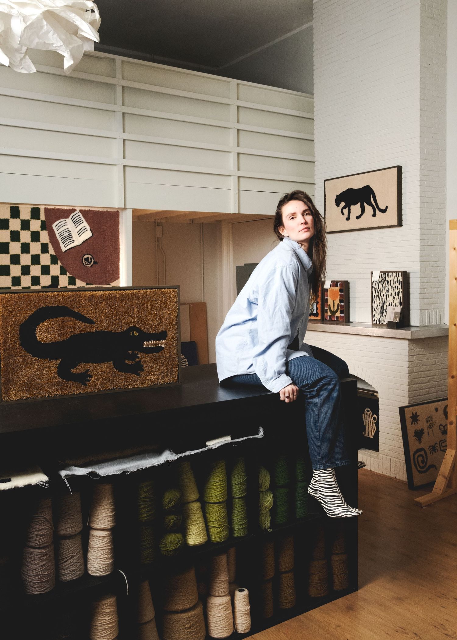 Woman sitting on a dark cabinet surrounded by yarn rolls and handmade rugs featuring animal motifs in a creative studio.
