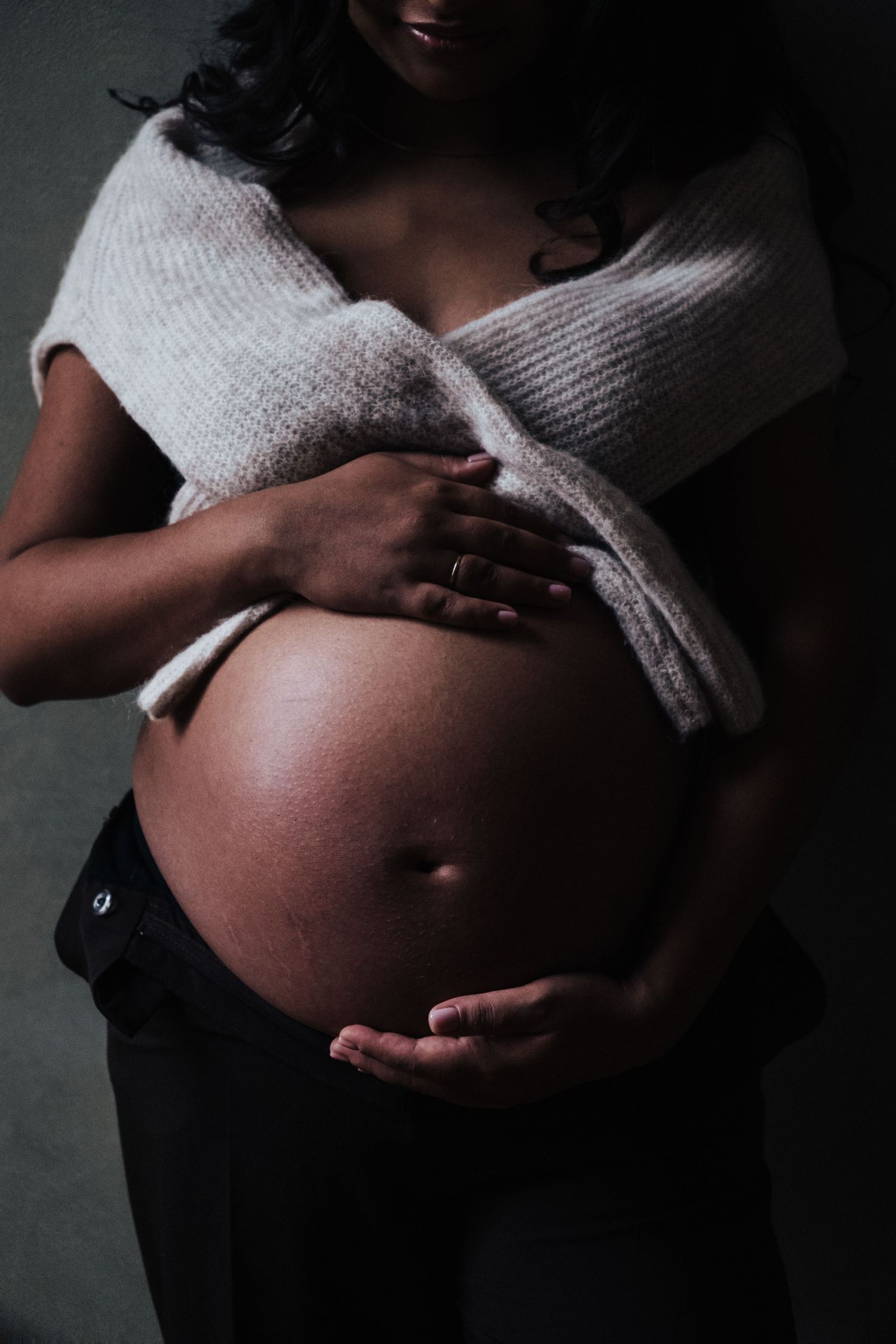 Intimate maternity photo showing pregnant belly wrapped in white sweater against dark background.