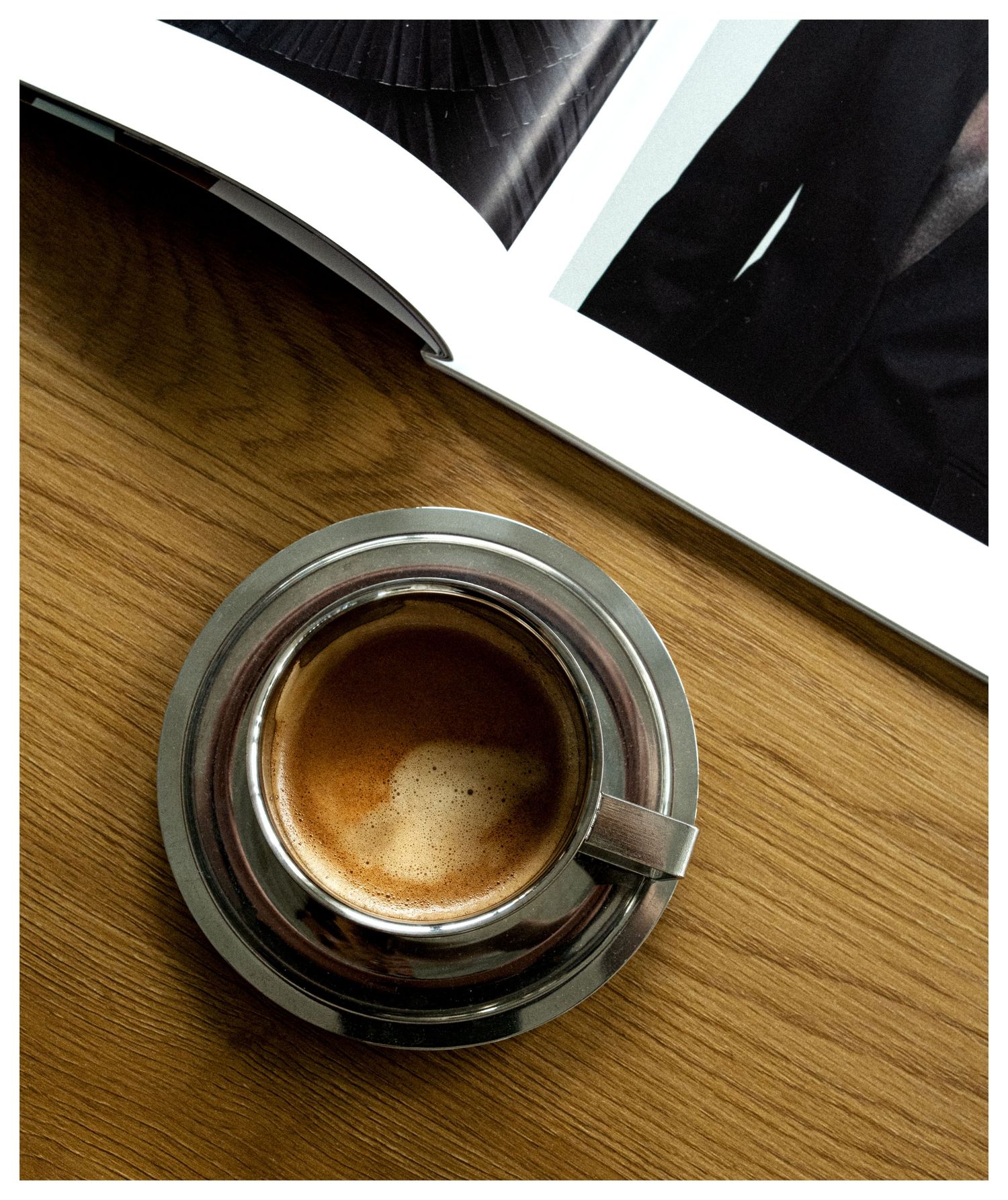 A steaming espresso in a silver cup and saucer on a wooden table beside an open magazine.
