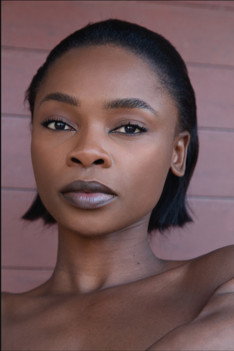 Close-up beauty portrait with sleek black hair, flawless skin, and matte mauve lipstick against a pink background.