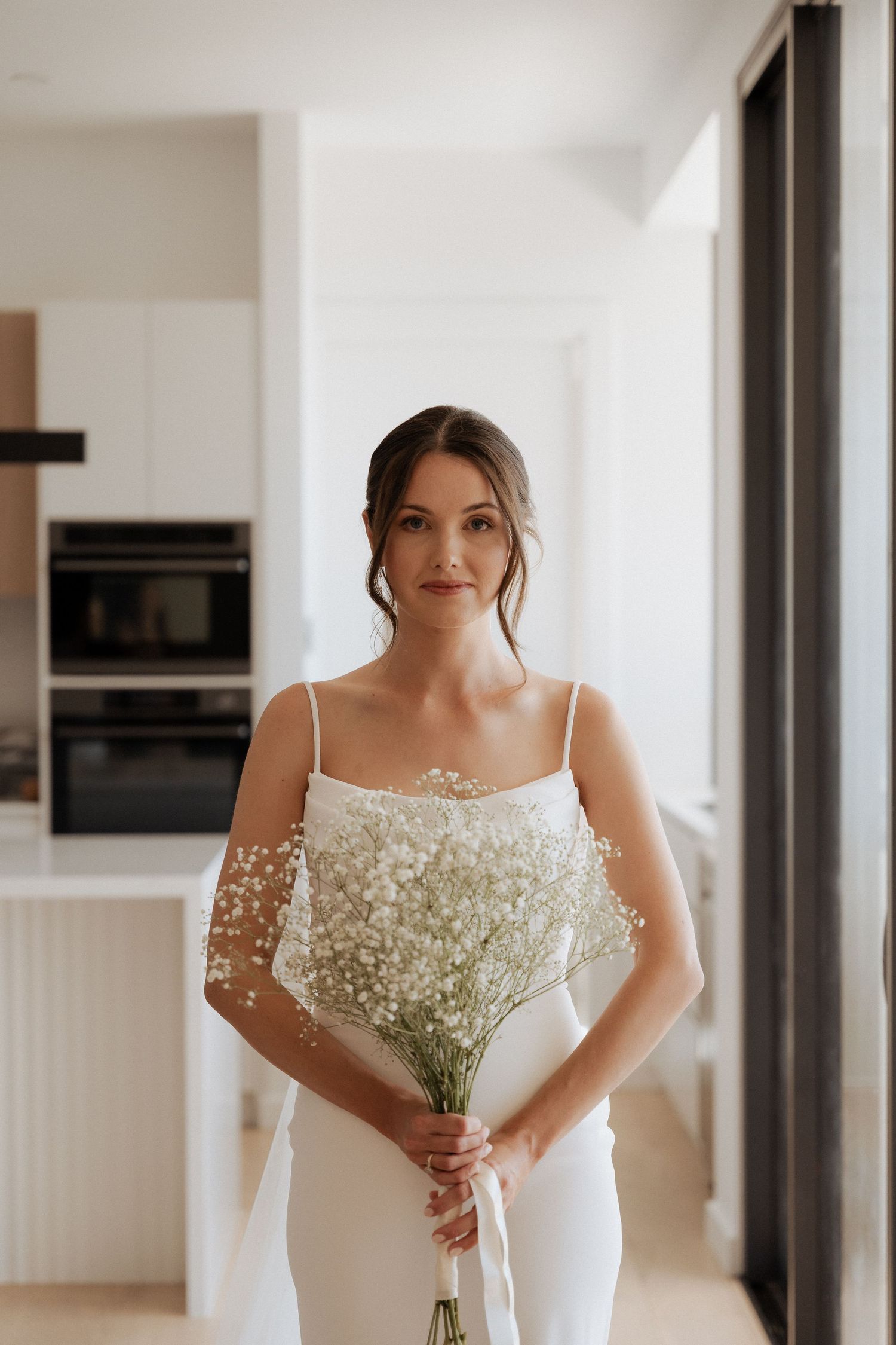 An elegant bride in a white spaghetti strap gown holds a delicate bouquet of baby's breath flowers.