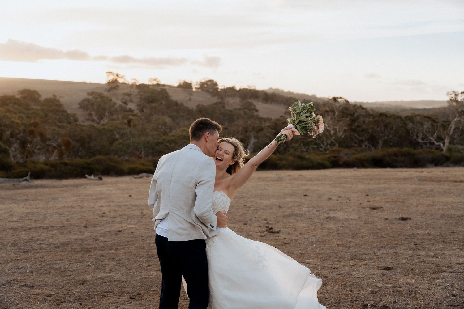 Wedding couple shares a playful dance move on an open field at sunset.