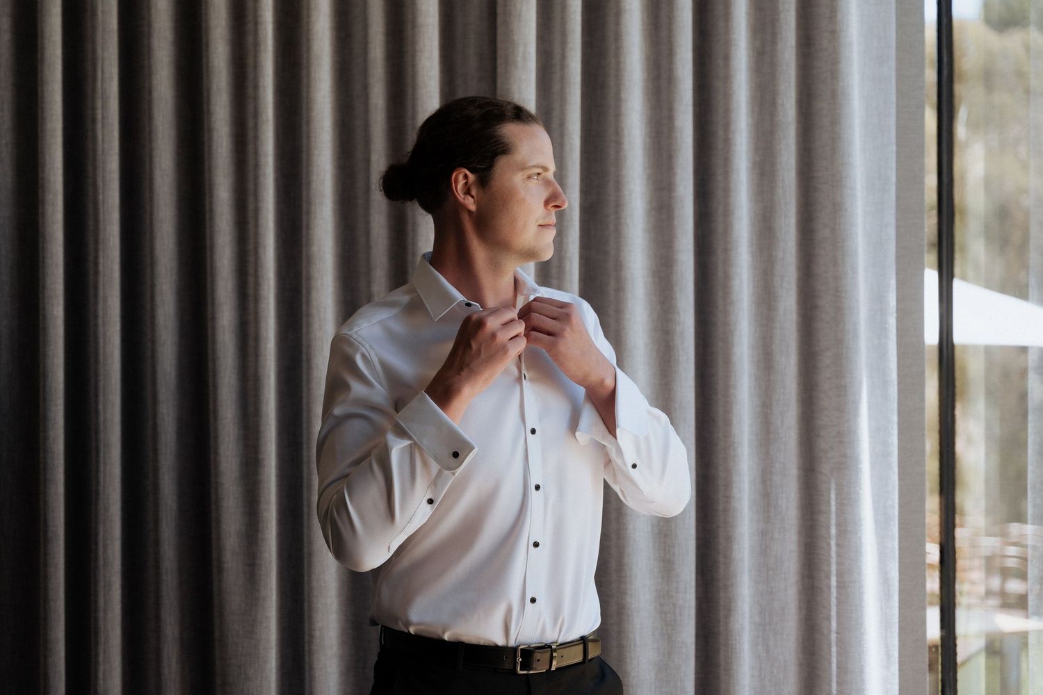 Person in formal white shirt adjusting tie while standing by curtained window. braytmedia adelaide weddings
