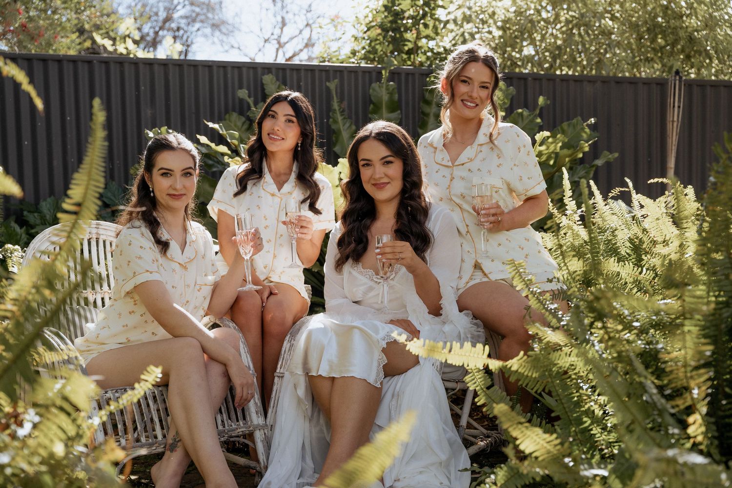 Group of women in white dresses pose together in a sunny garden setting for a bridal party photoshoot.